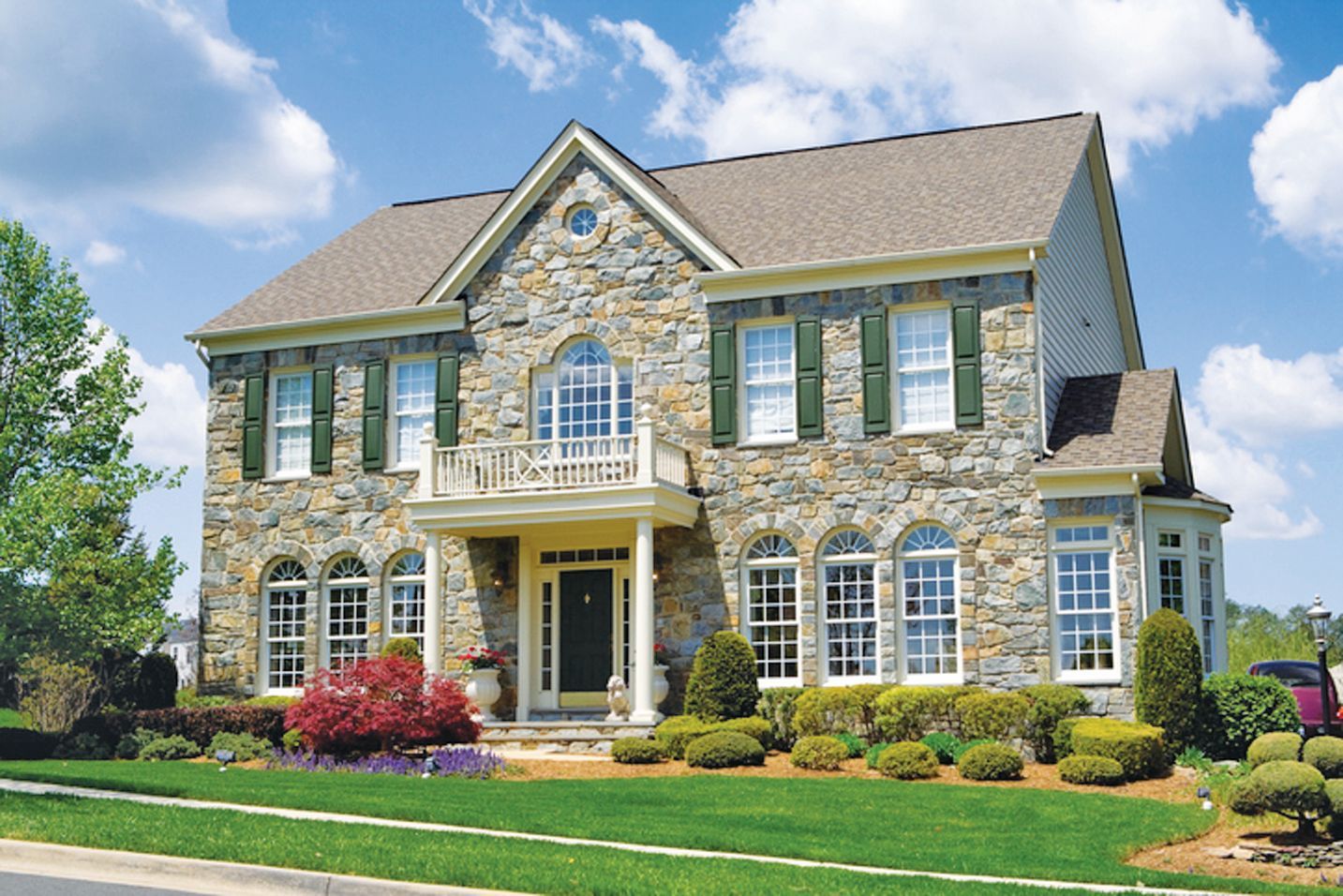 Two-story stone house with a gabled roof, a small front balcony, and a green lawn under a blue, cloudy sky.
