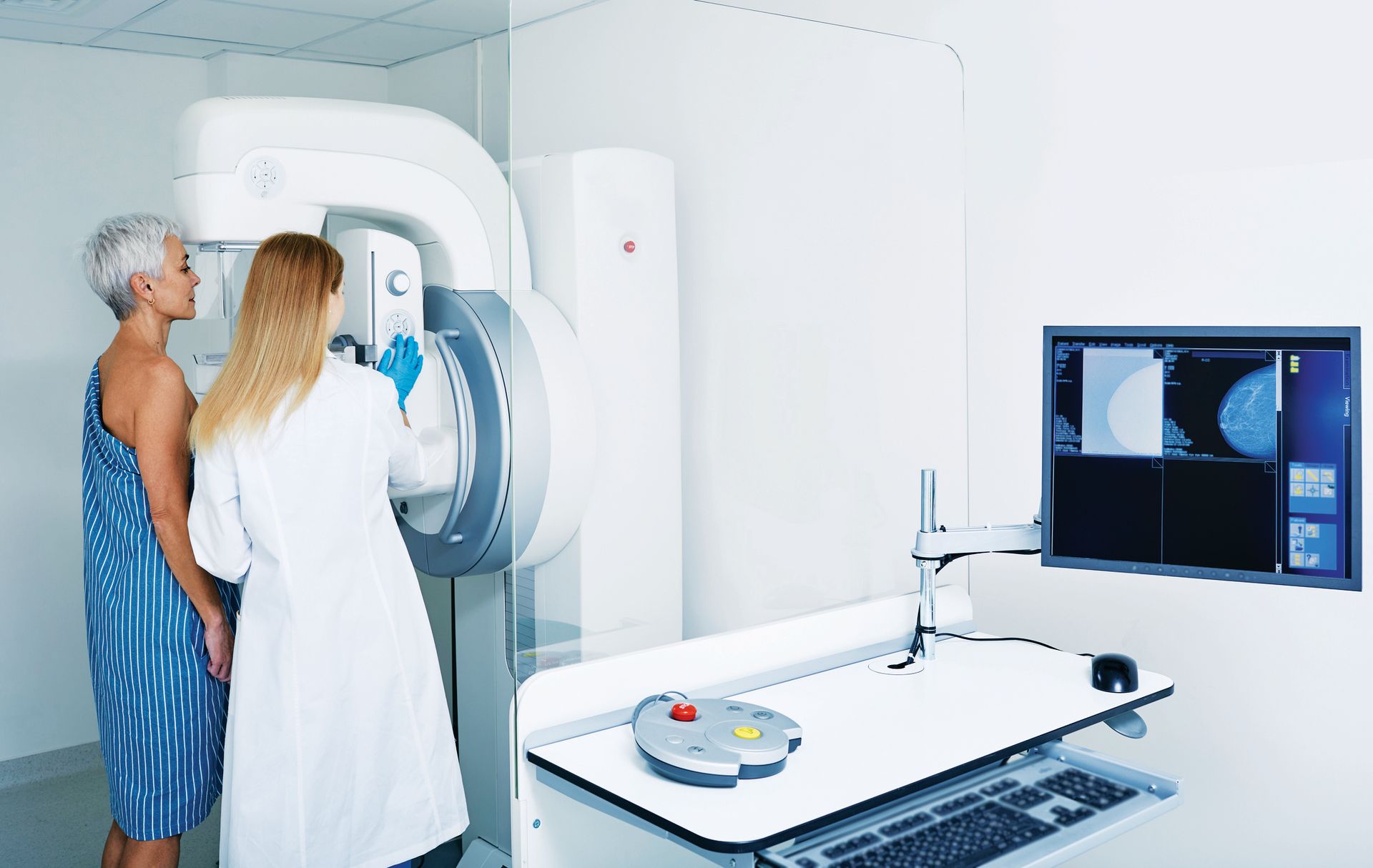 A technician in a white coat operates a mammography machine while a patient stands ready for an imaging procedure.