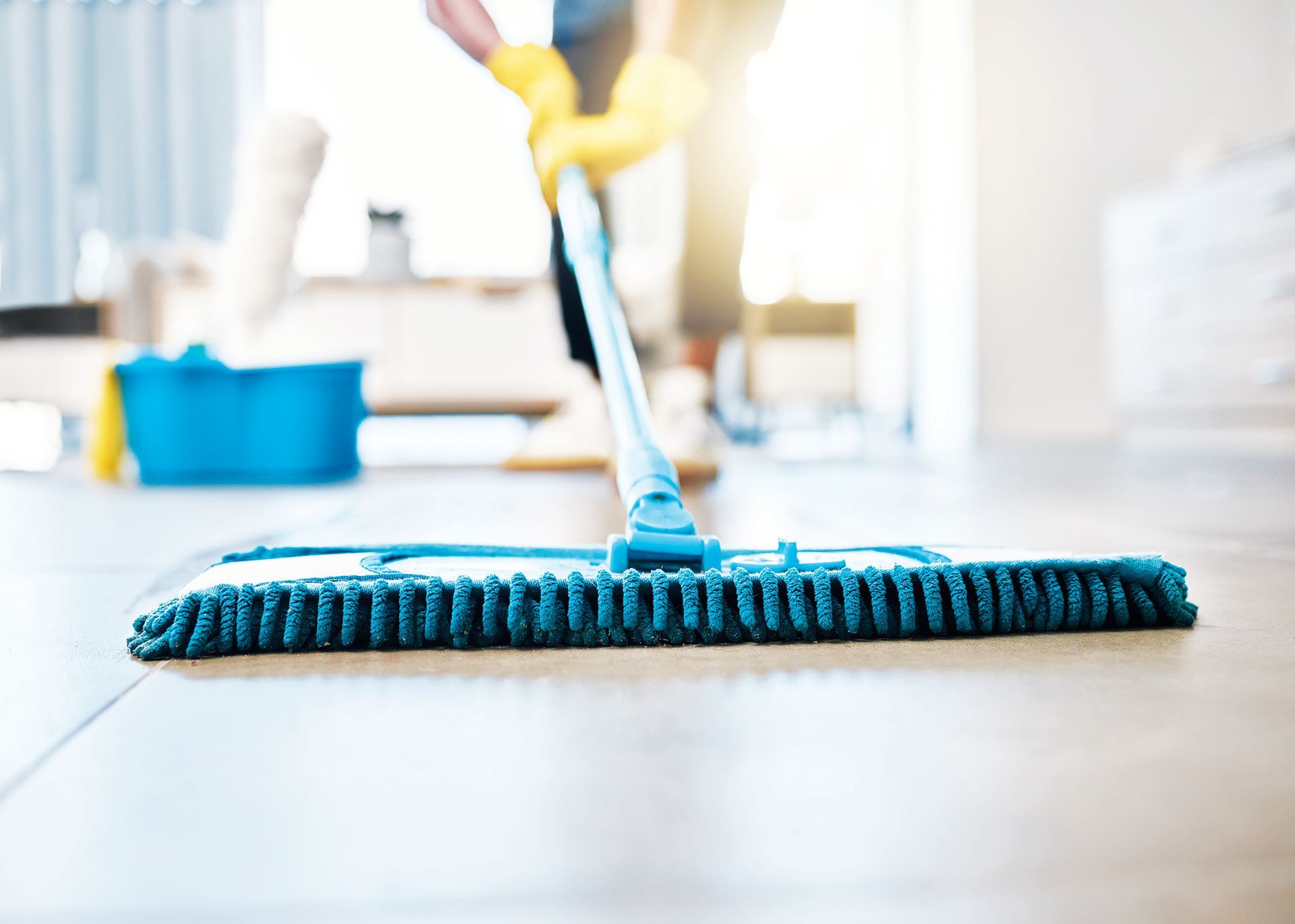Close-up of a blue mop cleaning a light-colored floor, with a person wearing yellow gloves pushing it in the background.