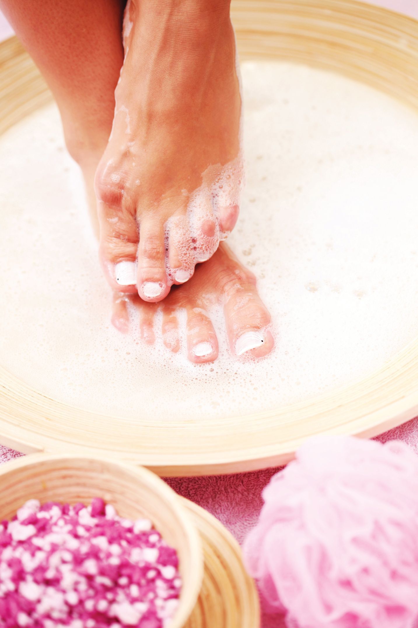 A pair of feet soaking in a wooden basin filled with bubbly water, next to a small bowl of pink bath salts and a puff.