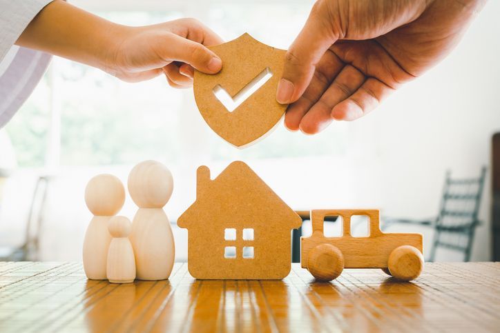 Hands hold a wooden shield with a check mark over wooden models of a family, a house, and a car on a wooden surface.