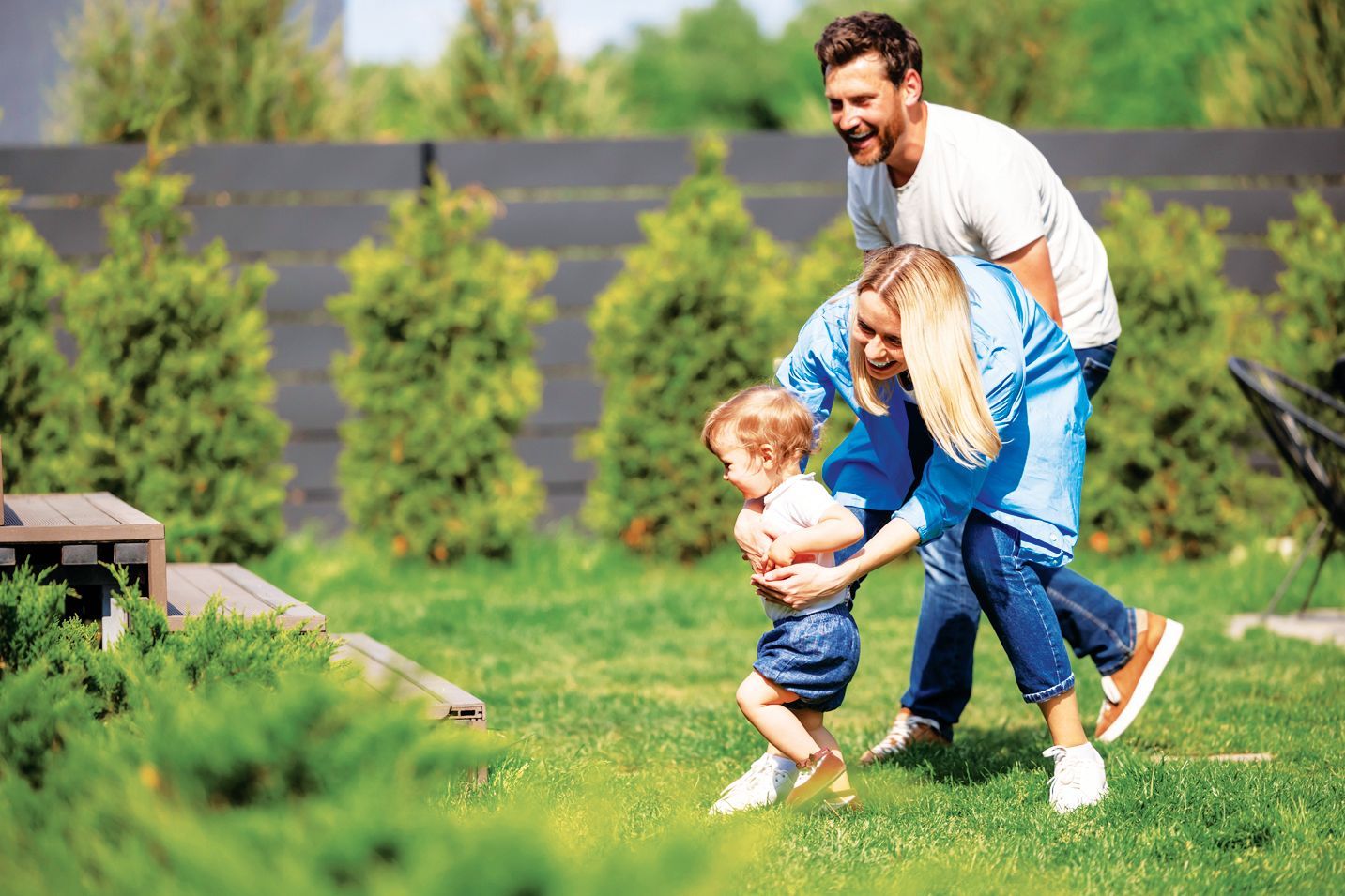 A parent walks behind a small child in a grassy backyard while another adult leans down to help them walk.