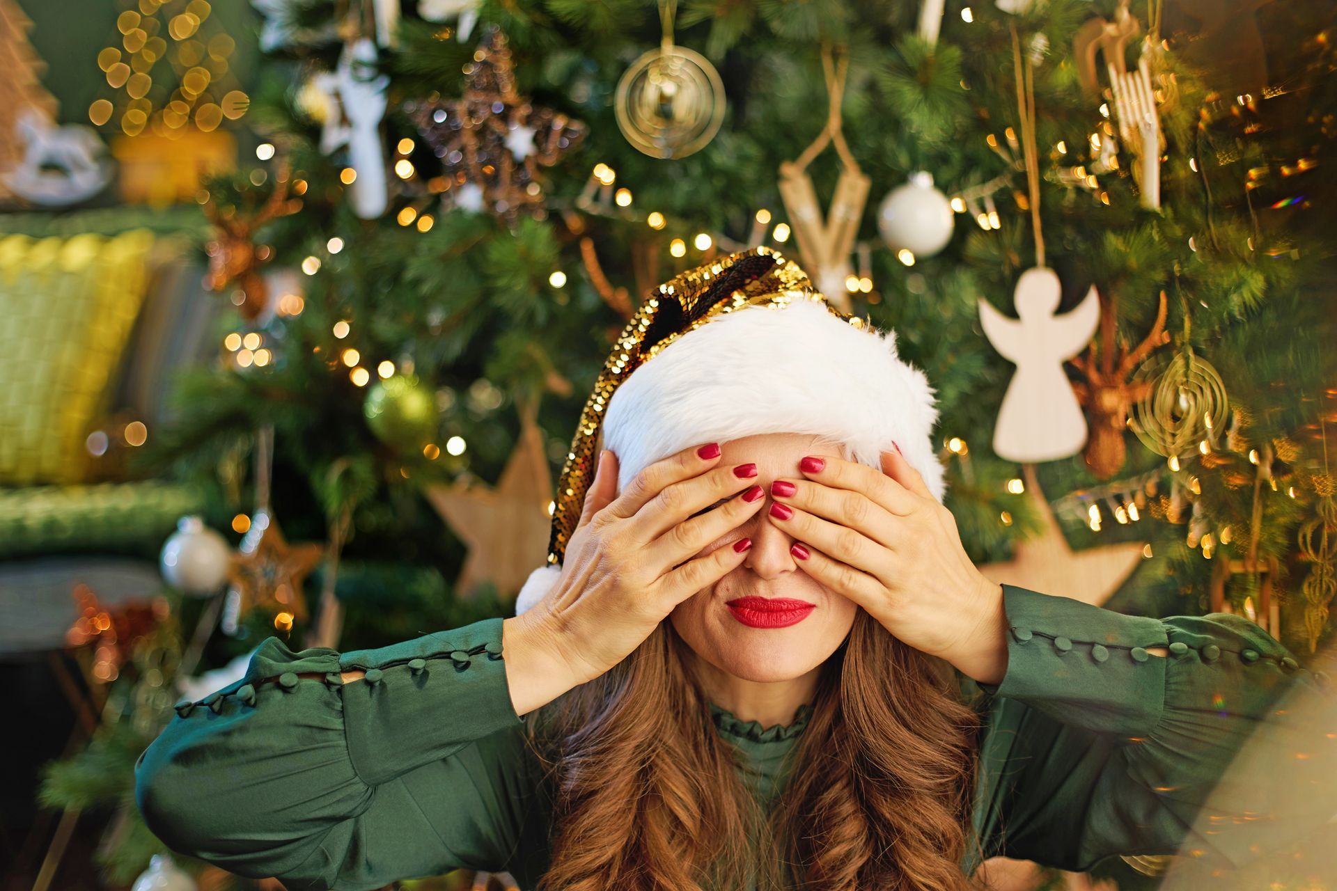 A person wearing a Santa hat and green top, with hands covering their eyes, posed in front of a decorated Christmas tree.