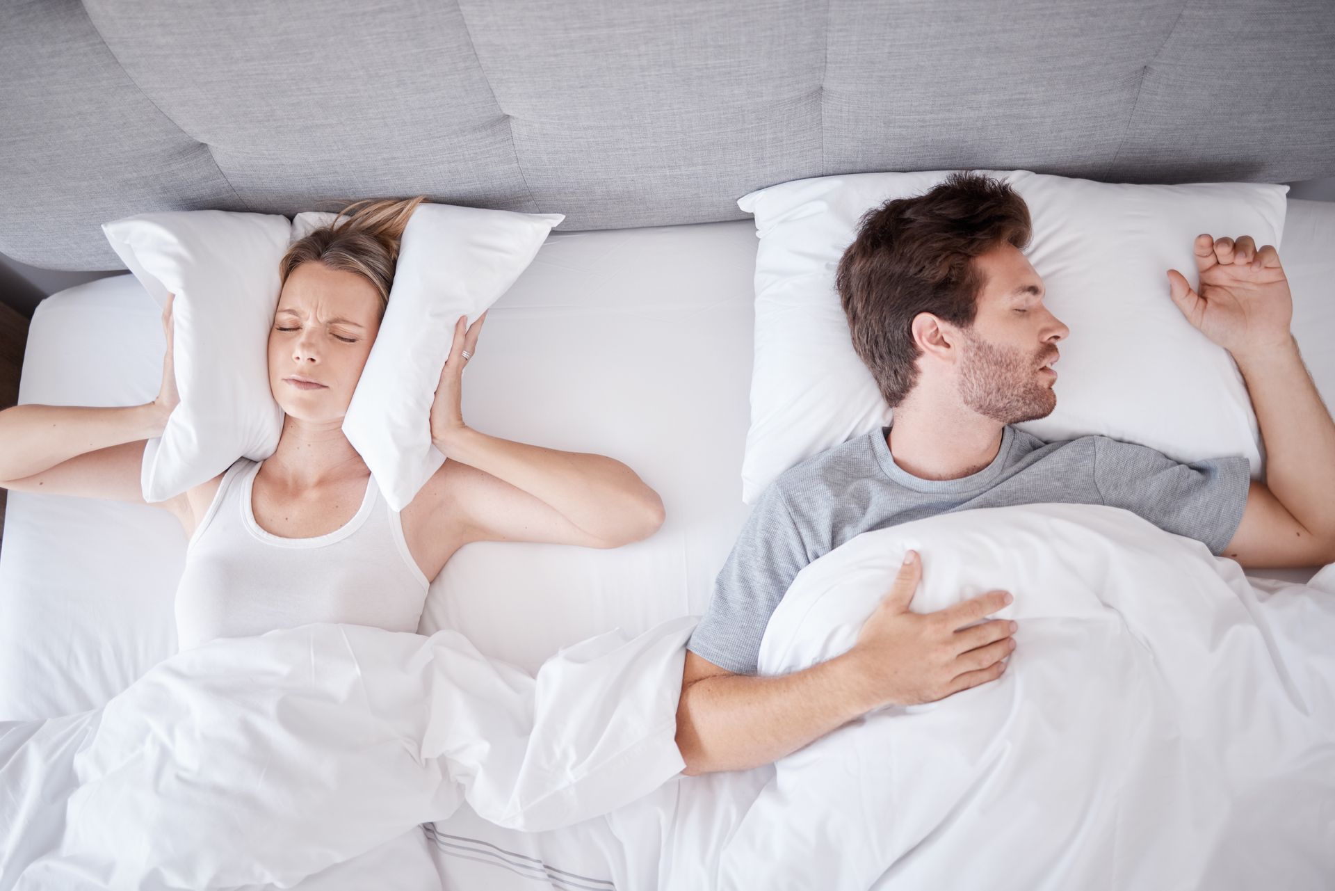 A person in bed covers their ears with a pillow in frustration while their sleeping partner rests peacefully nearby.