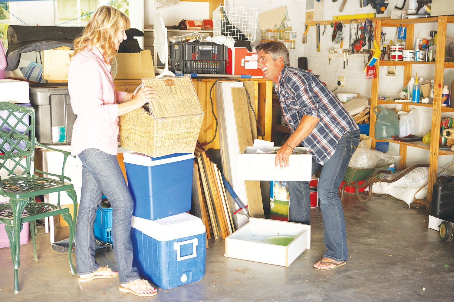 A woman and a man organize items in a cluttered garage, holding boxes and coolers while standing on a concrete floor.