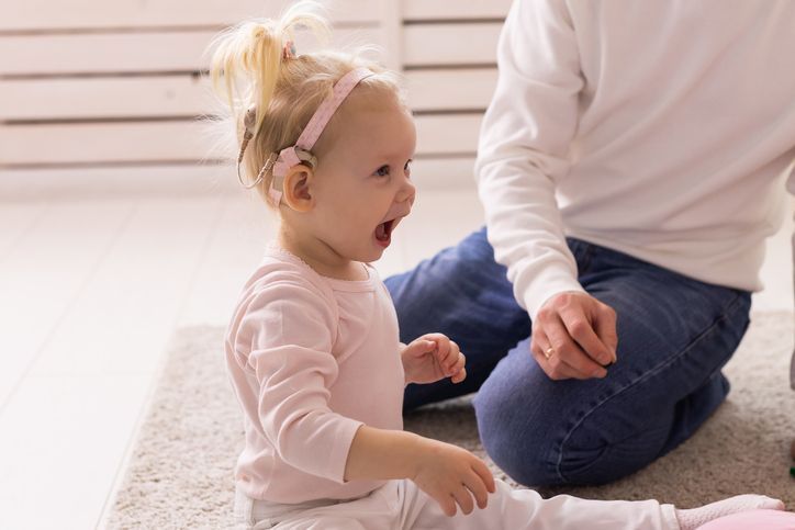 A toddler with a cochlear implant laughs while sitting on a rug near an adult.