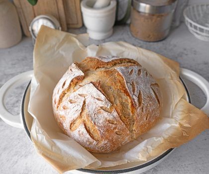A freshly baked, golden-brown loaf of sourdough bread with decorative scoring, resting on parchment paper in a Dutch oven.