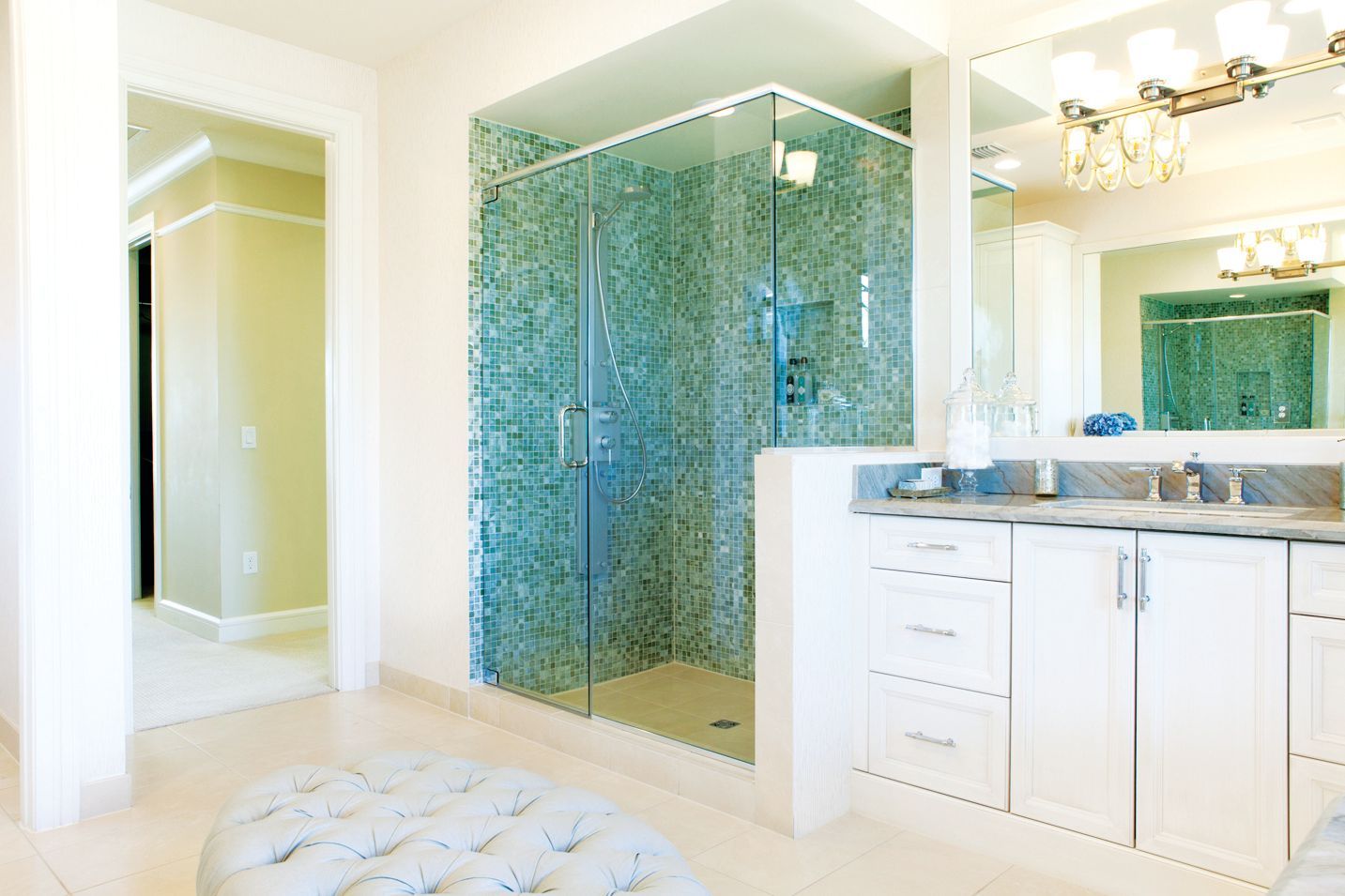 A bright bathroom with a glass-enclosed shower featuring turquoise mosaic tiles, a white vanity, and a tufted ottoman.