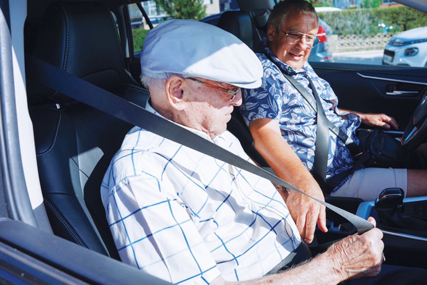 Two people sit in the front seats of a car, both wearing seatbelts and looking down while adjusting their safety straps.