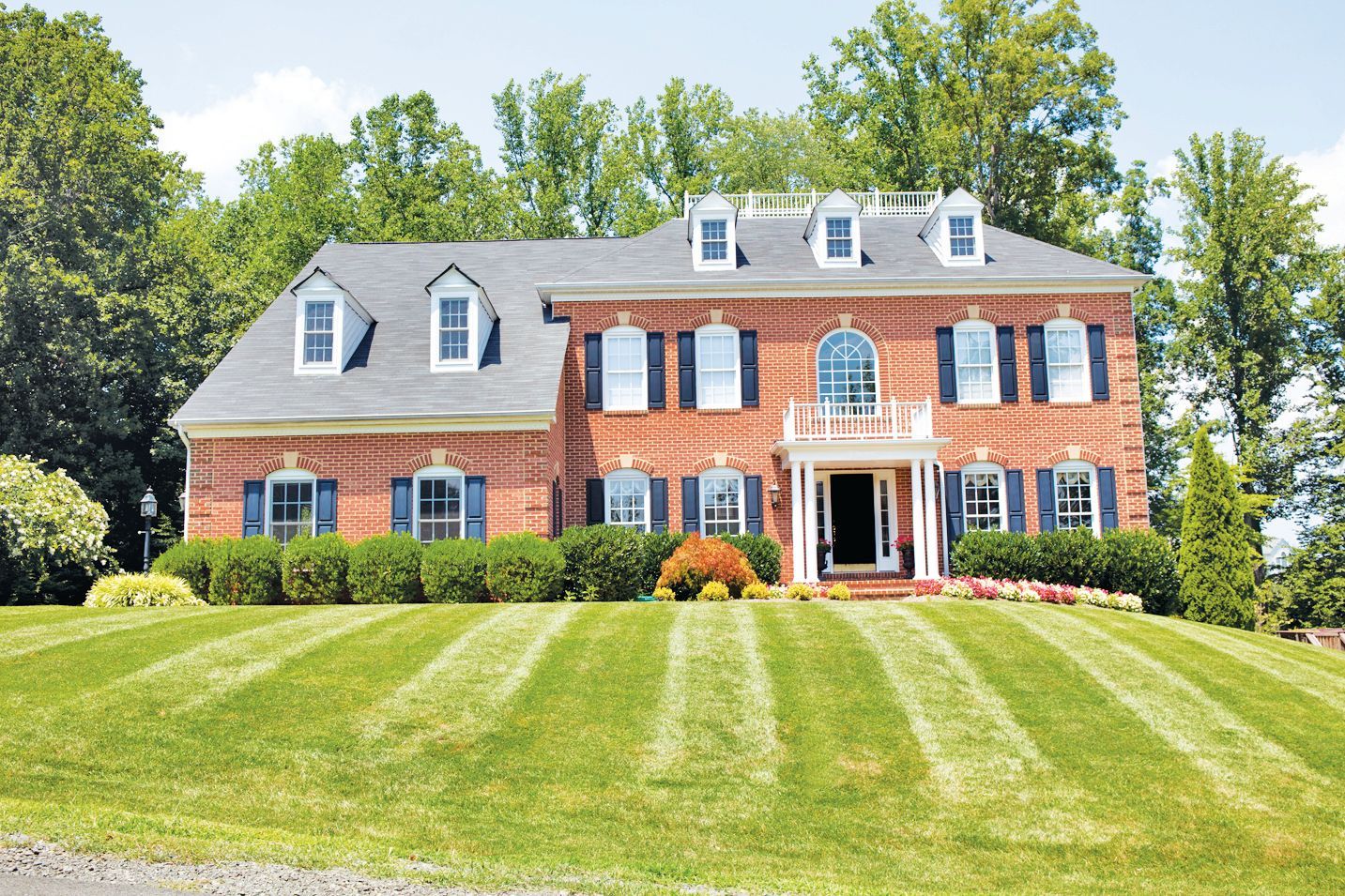 A two-story red brick colonial house with a striped lawn, gabled dormers, and a white front porch.