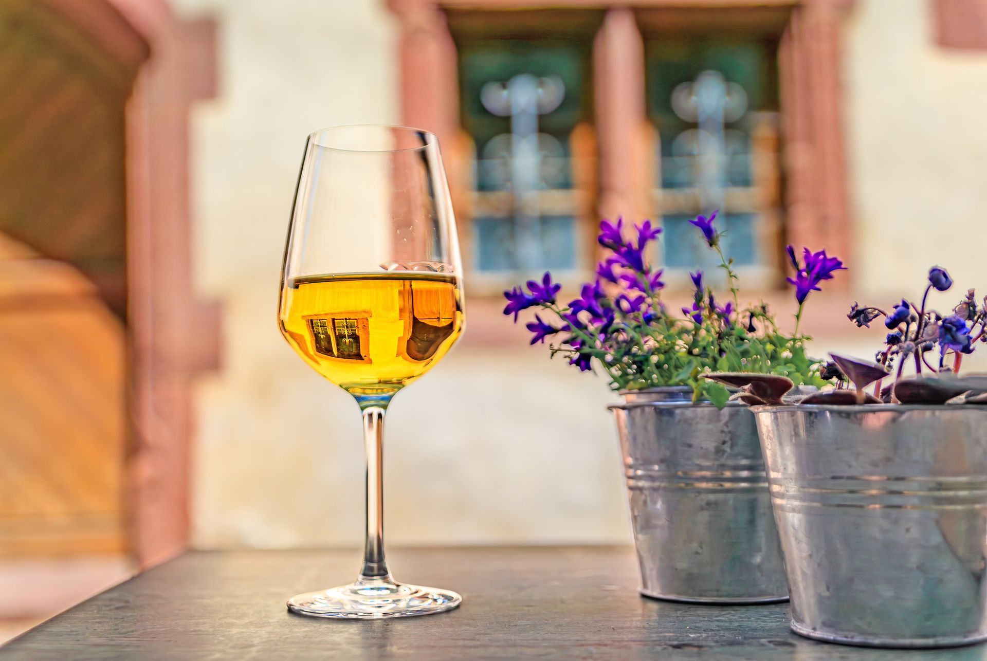 A glass of golden white wine next to two small silver buckets of purple flowers on a table outdoors.