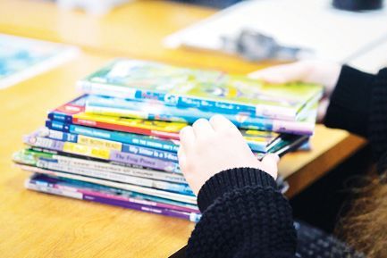 A person in a black sweater holds a stack of colorful children's books on a light wood table.