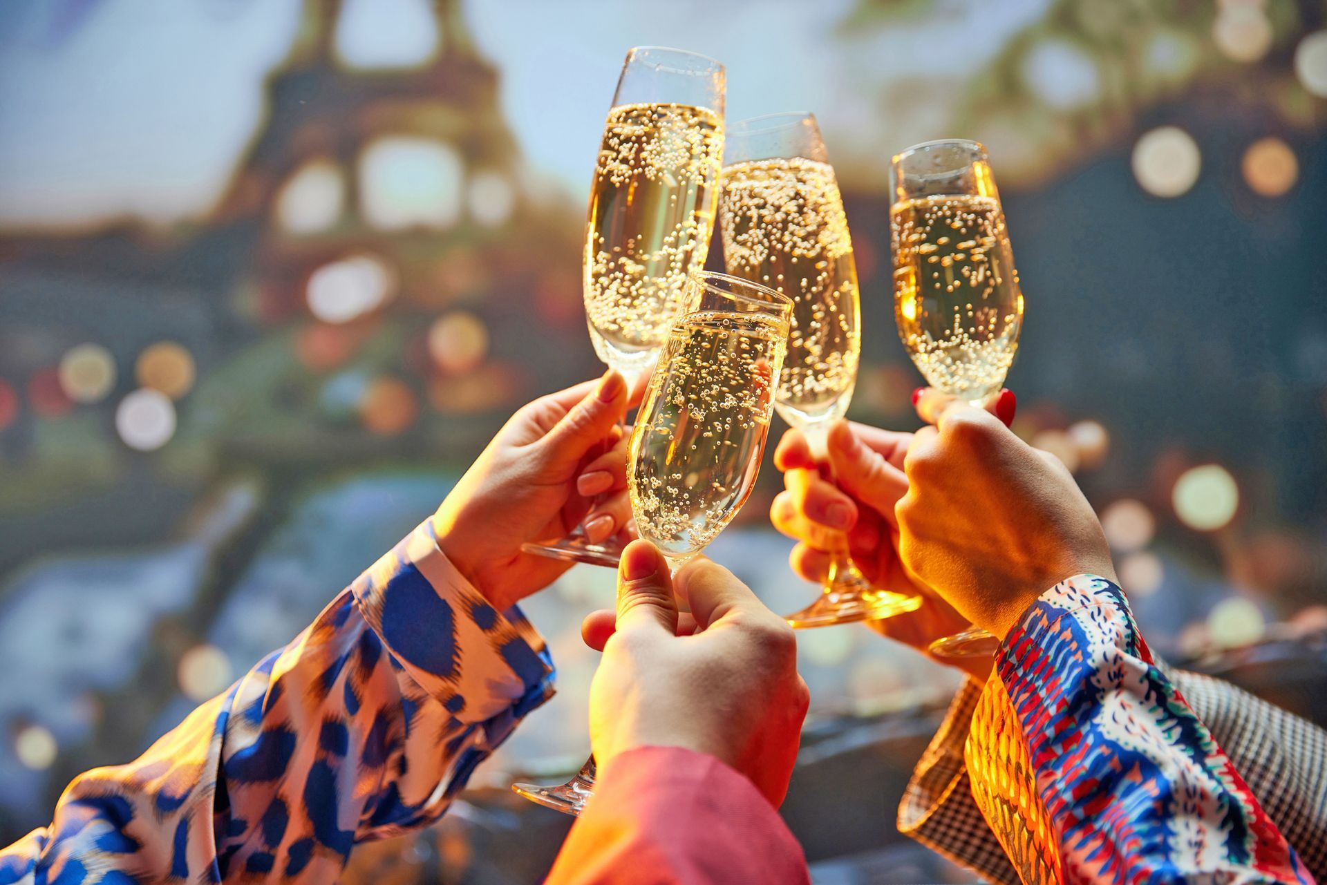 Hands holding four champagne flutes for a toast against a blurred background featuring the Eiffel Tower at night.
