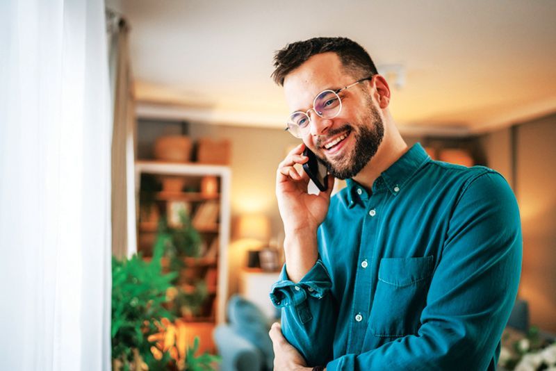 A person with a beard and glasses wears a teal shirt, smiling while talking on a smartphone indoors.