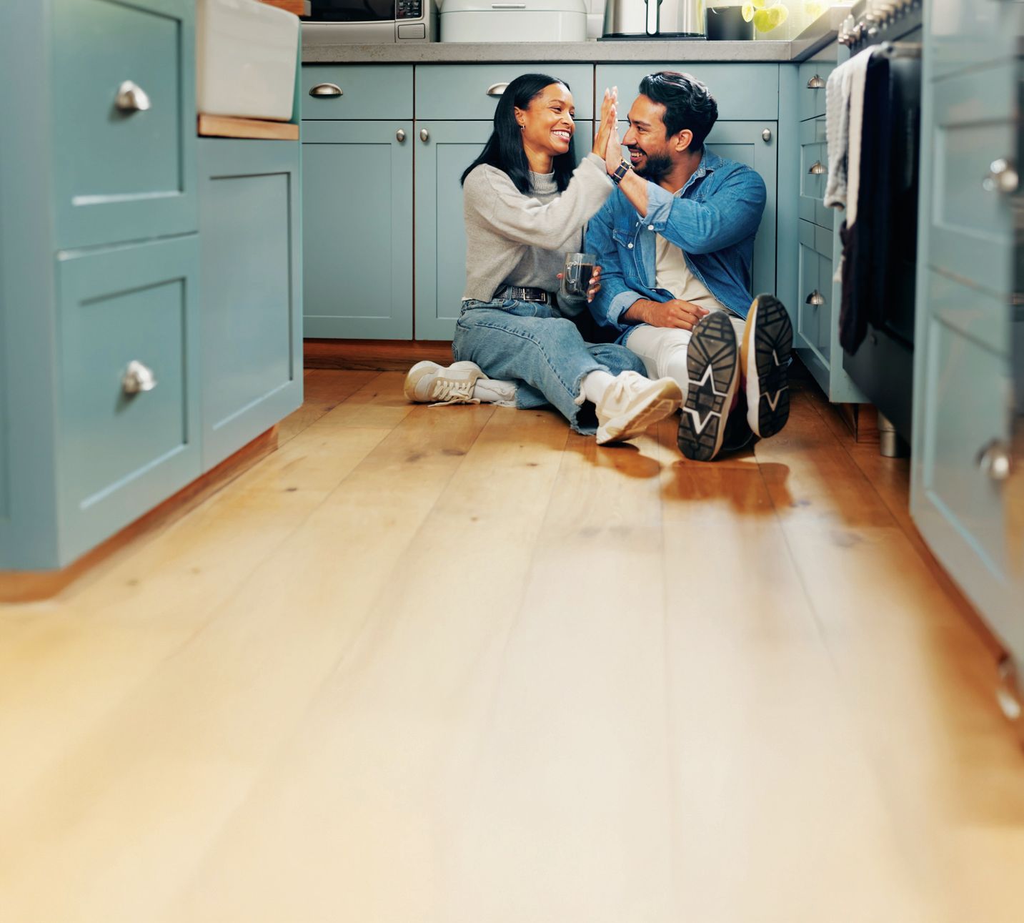 Two people sit on a wooden kitchen floor, smiling and giving each other a high-five in front of blue cabinets.