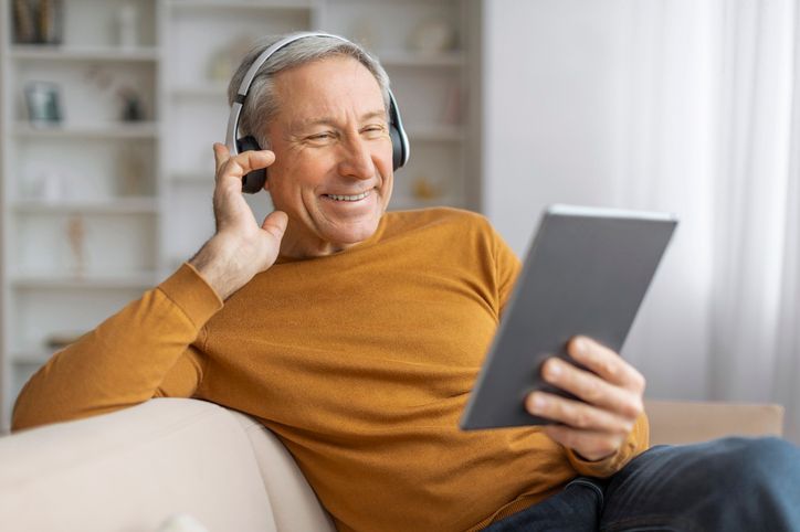 A person in an orange sweater wearing headphones, smiling while looking at a tablet while sitting on a couch.
