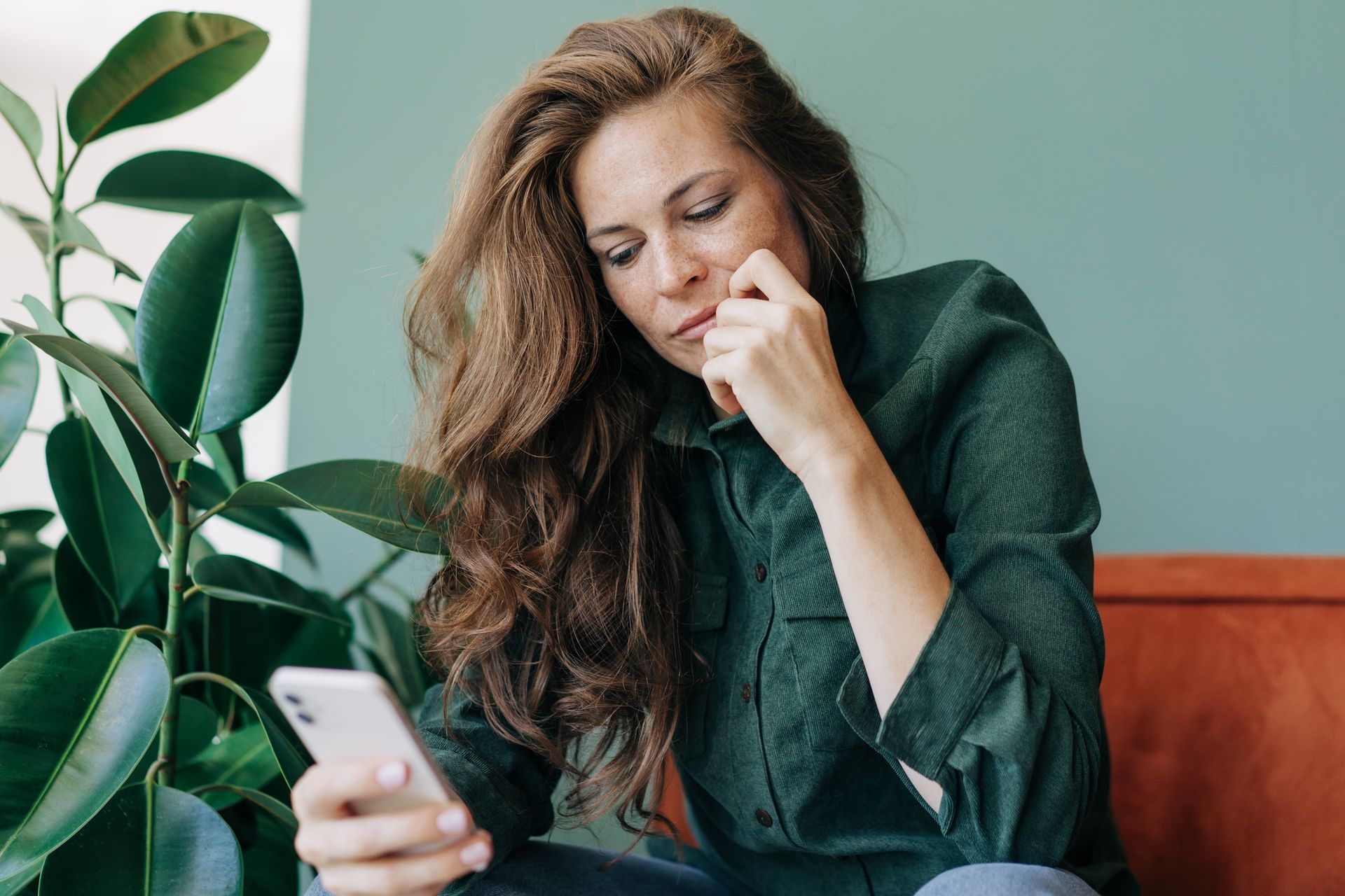 A person with long, wavy brown hair wearing a dark green shirt sits by a leafy plant and looks at a smartphone.