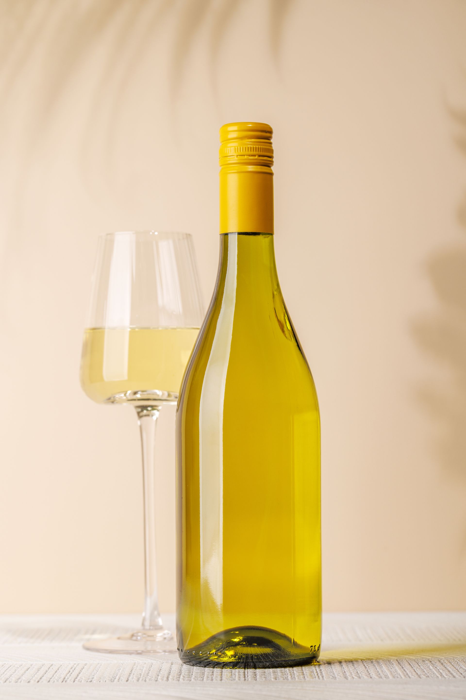 A bottle of white wine with a yellow cap and a filled wine glass, set against a soft, beige background with palm shadows.