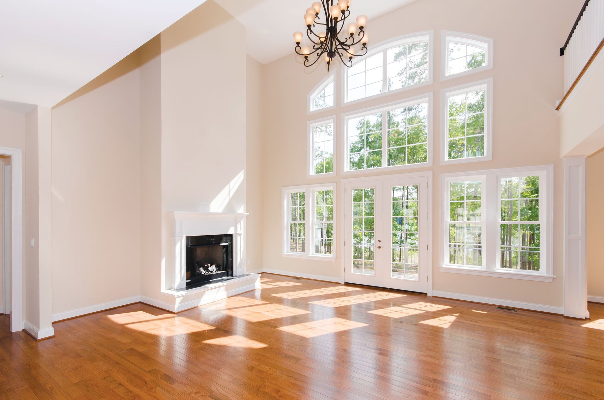 Empty, sunlit living room with high ceilings, hardwood floors, a fireplace, and large windows with a view of trees.