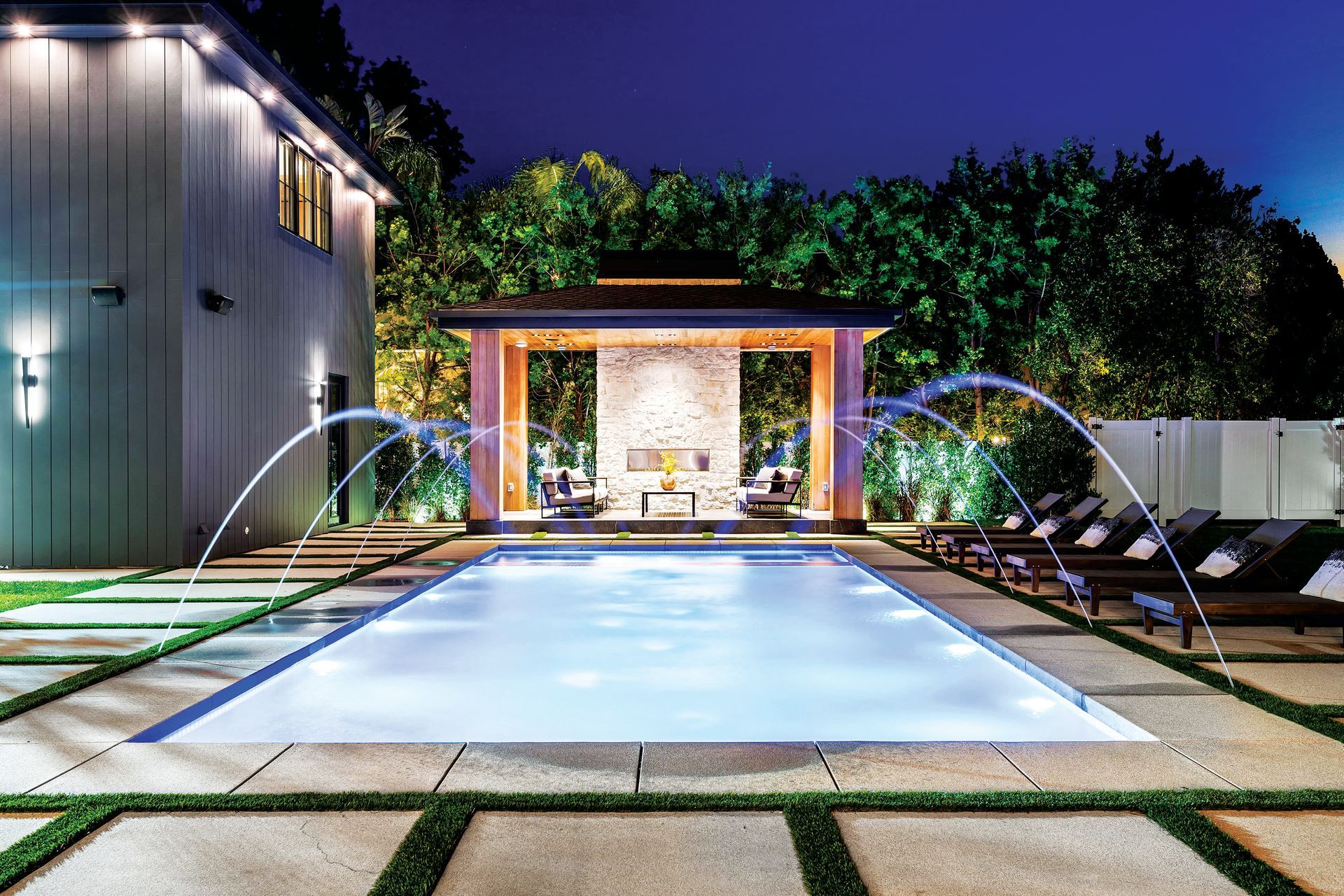 A lit, rectangular swimming pool with fountains, adjacent to a patio, lounge chairs, and a covered fireplace at night.
