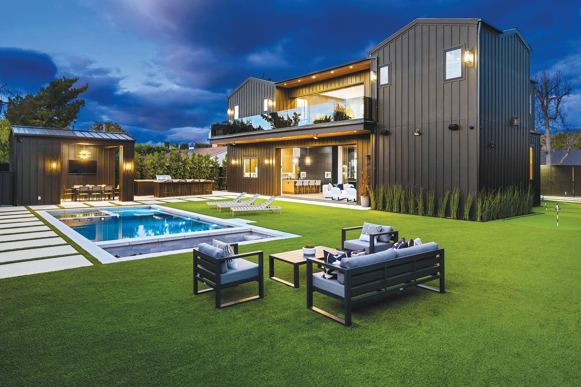 A modern dark-paneled two-story house with a swimming pool, lawn furniture, and an illuminated outdoor patio at twilight.