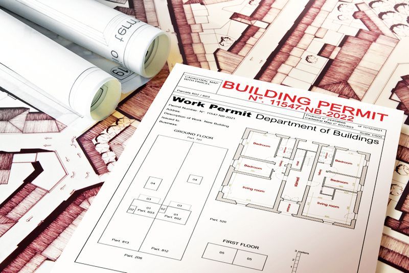 A printed building permit document rests on top of rolled architectural blueprints and a large site map.