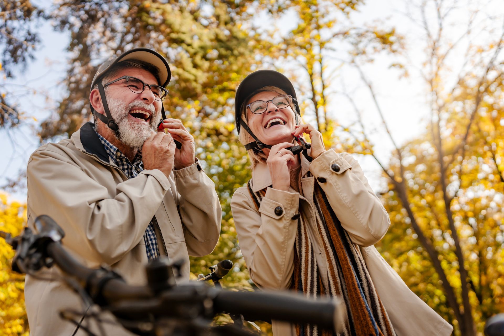 Two people laughing while putting on bicycle helmets in a park during autumn.