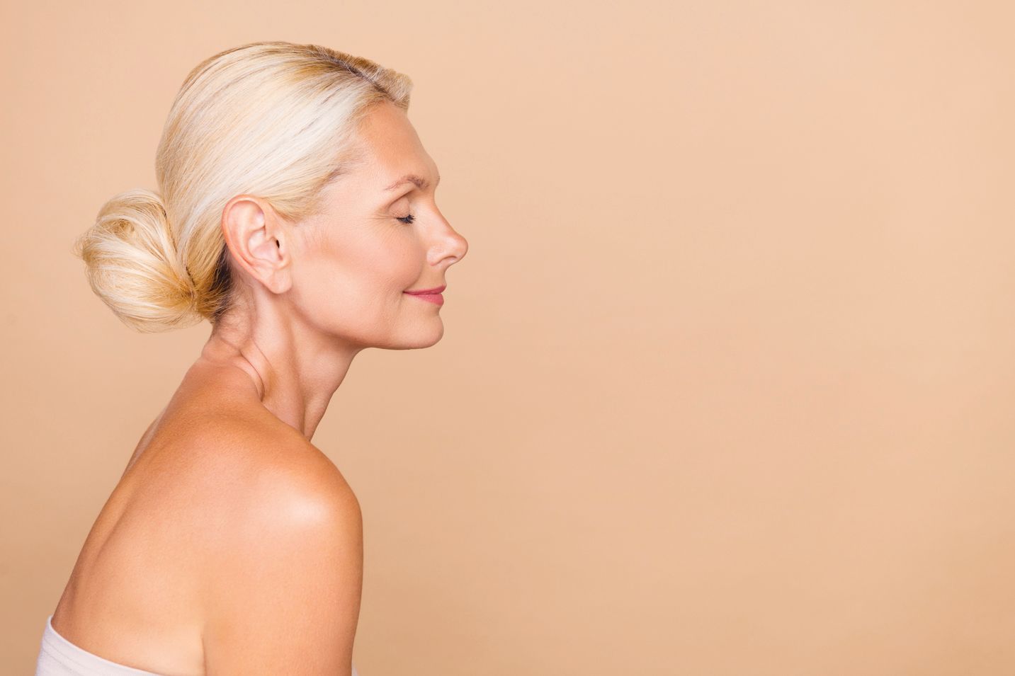 A person with blonde hair in a bun stands in profile against a solid beige background, smiling with eyes closed.