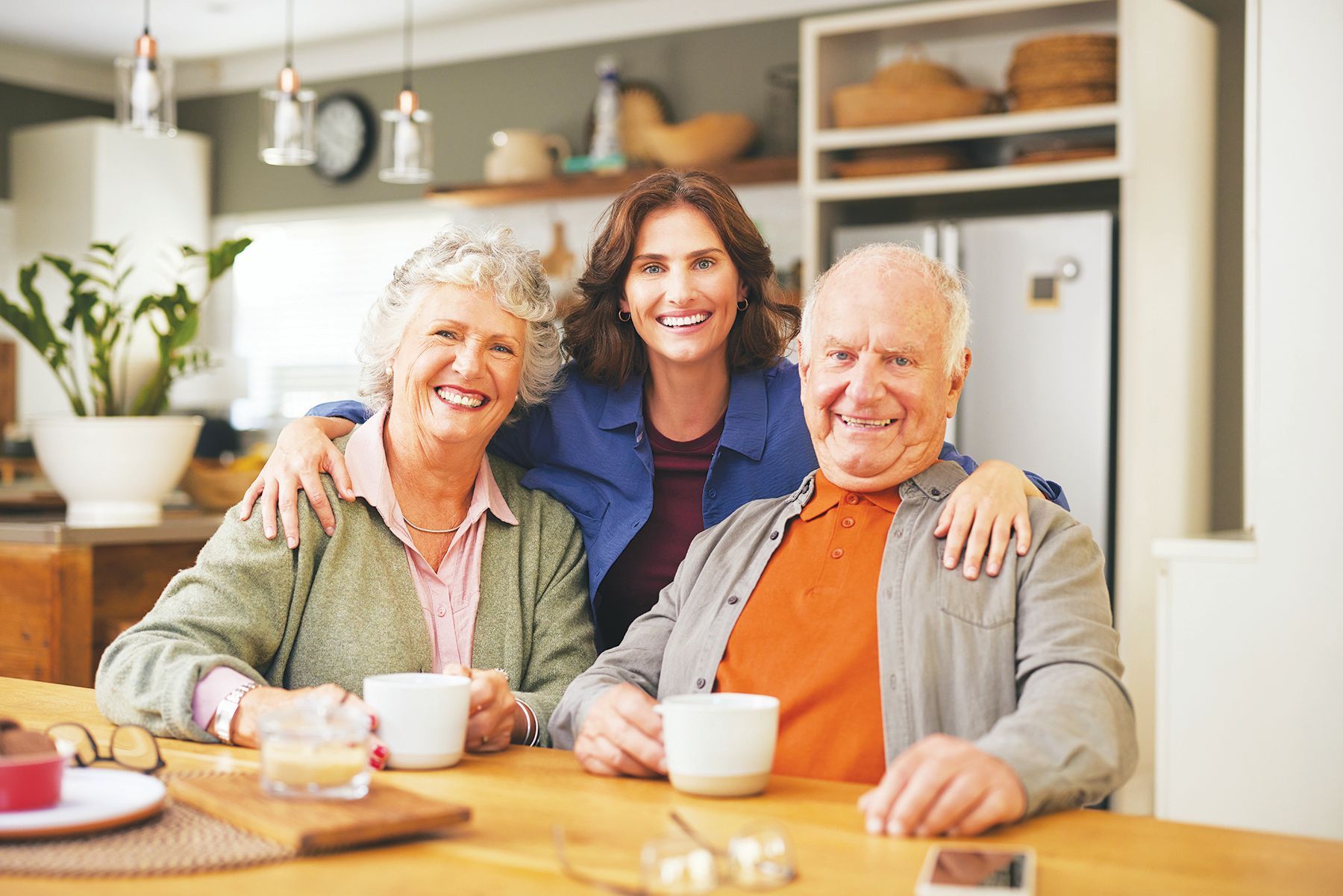 Three people smile while sitting at a wooden table in a bright kitchen with mugs in front of them.
