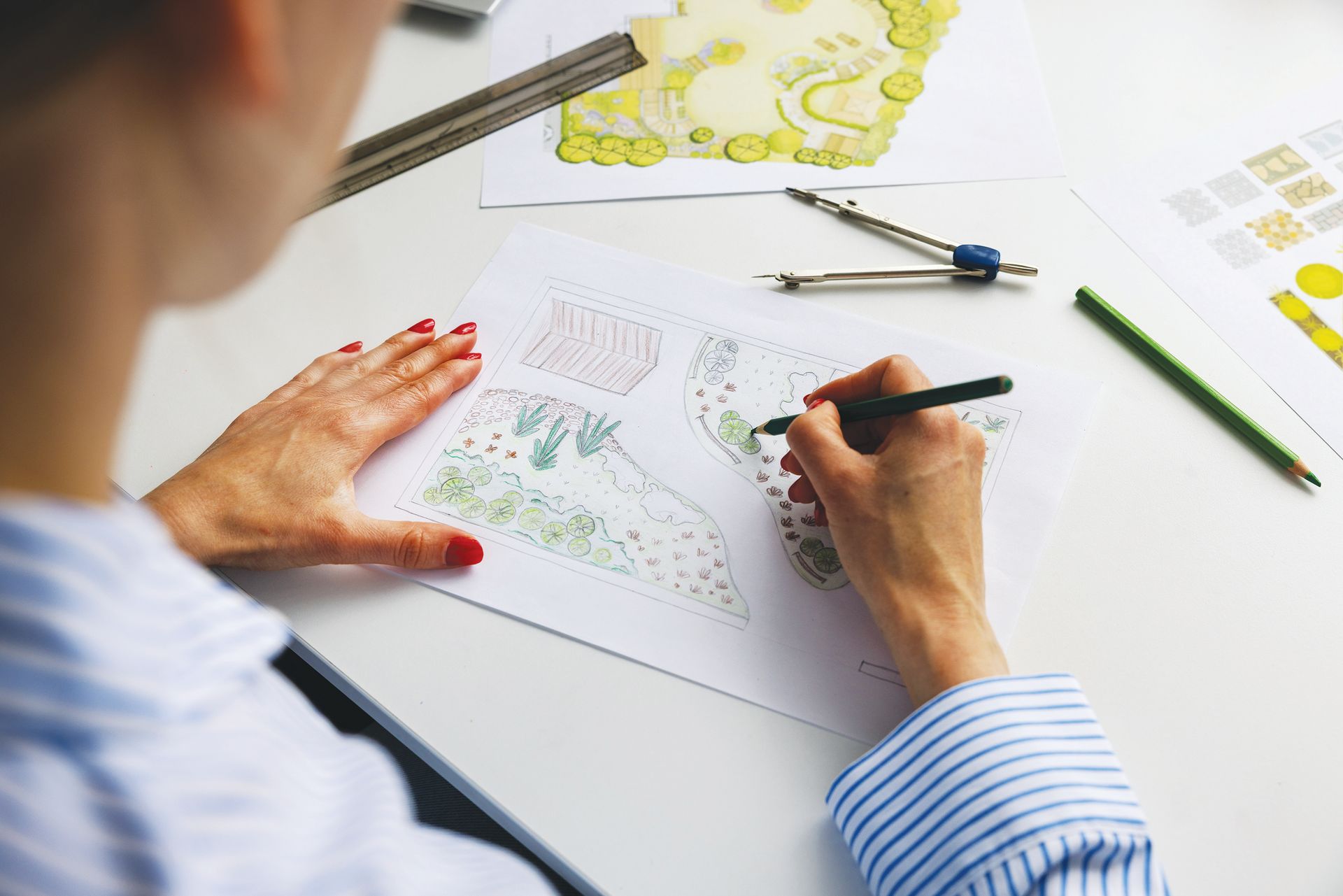 A designer in a blue striped shirt sketches a landscape plan on paper, surrounded by drawing tools on a white desk.