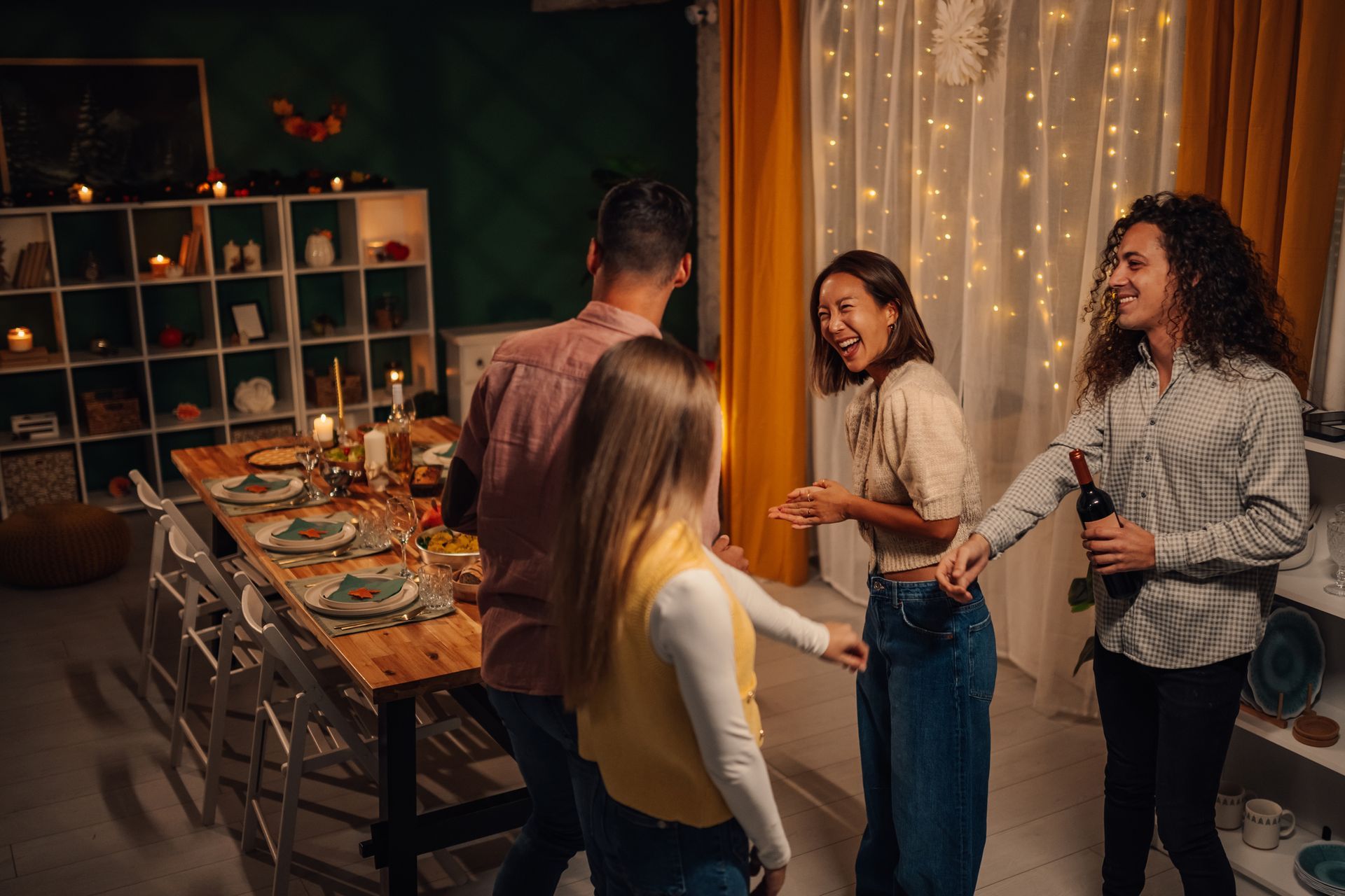 Four people socialize and laugh in a warmly lit dining room with table settings, fairy lights, and open shelving.