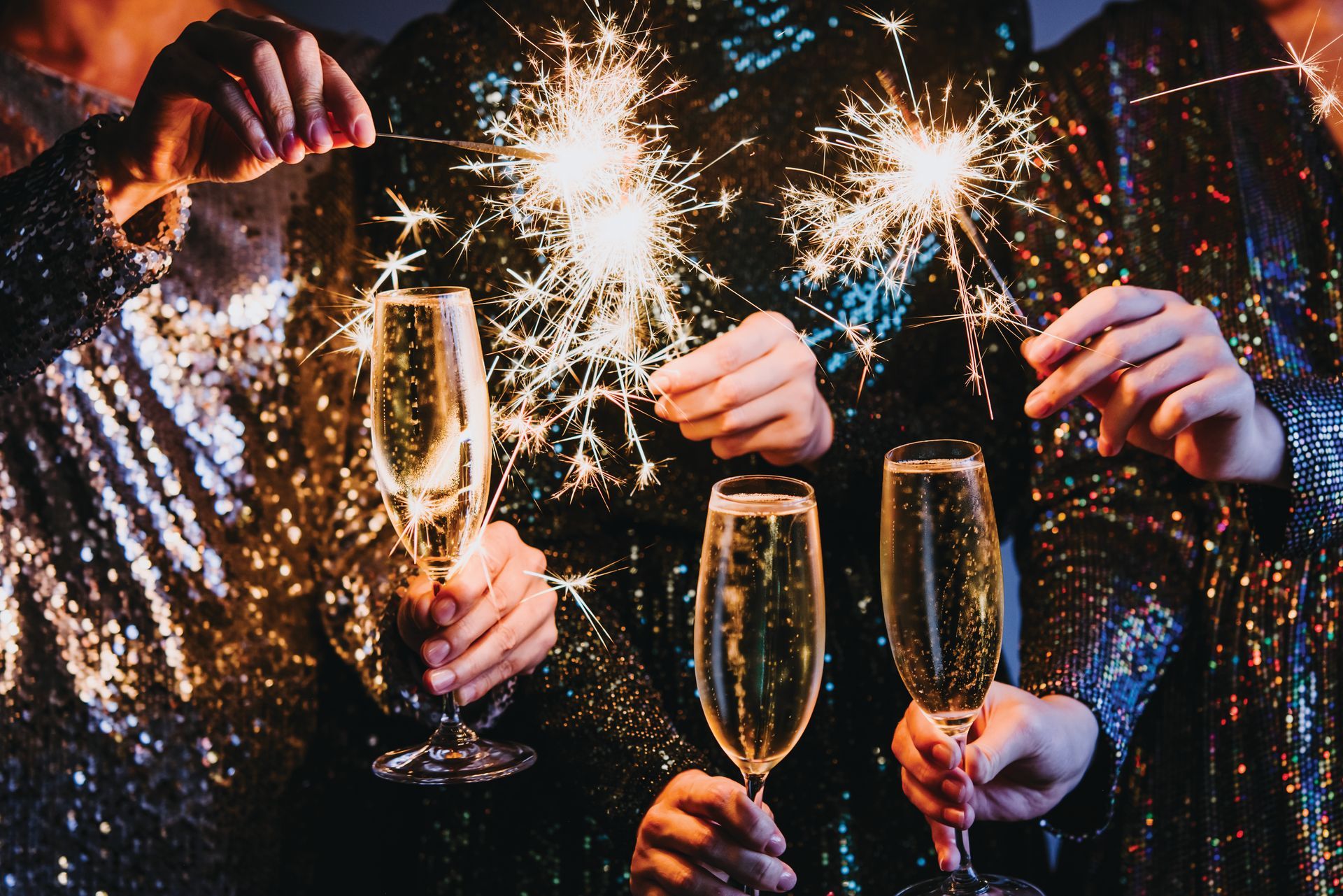 People in shimmering outfits hold champagne flutes and lit sparklers in a festive, dark-lit celebration.