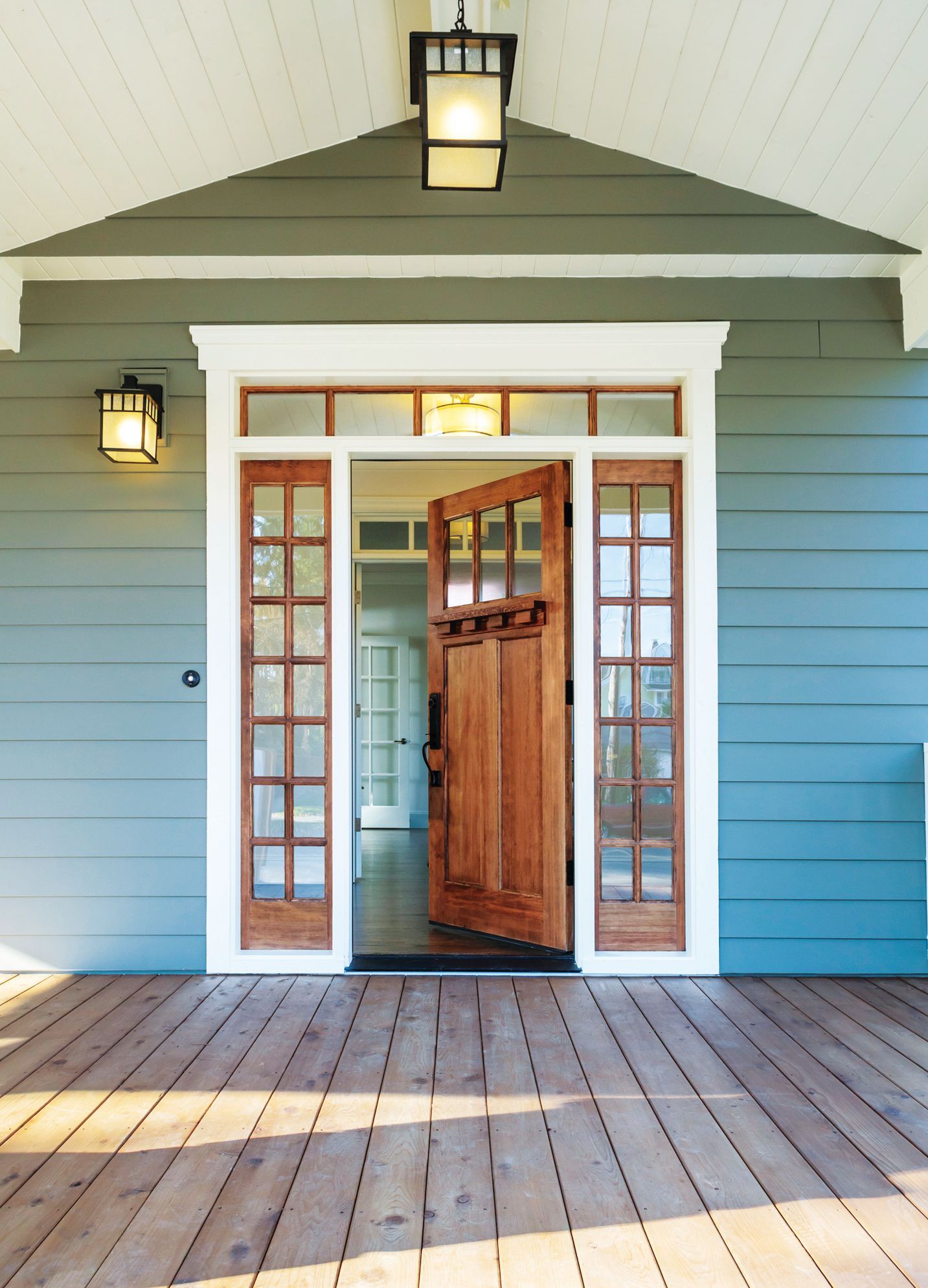 A wooden front door with side windows and a transom, set in a blue-sided house with a wooden porch floor and lanterns.