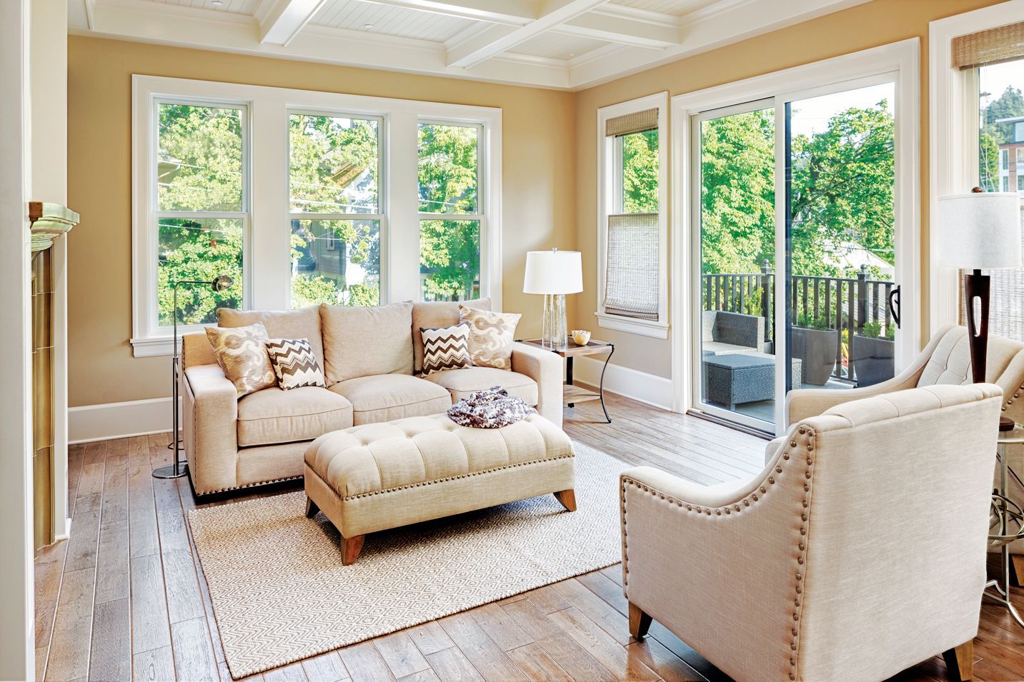 A bright living room with beige sofas, a tufted ottoman, and a patterned area rug on wood floors, facing large windows.