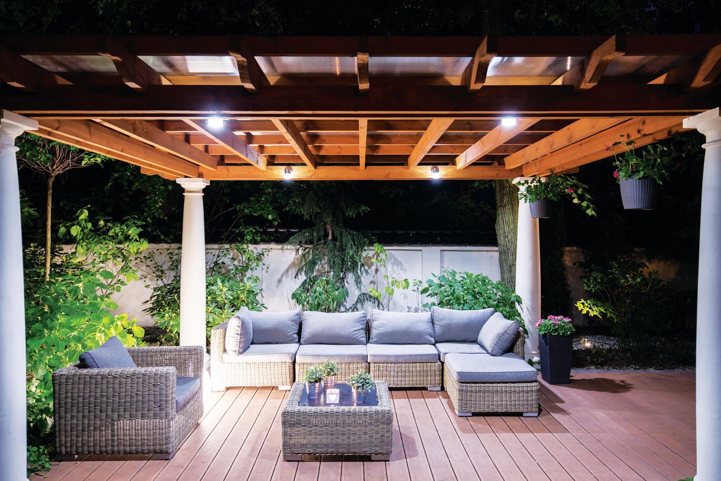 An outdoor wooden pergola at night with a cozy wicker sectional, armchair, and coffee table set on a patio.