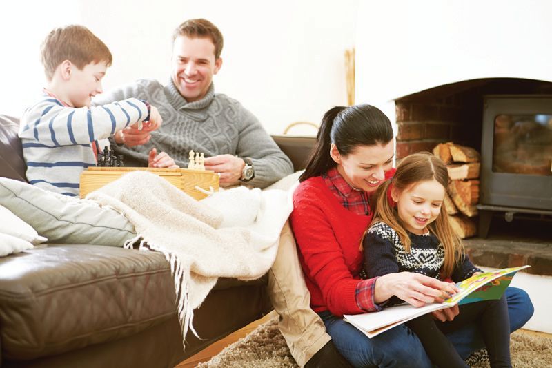 A family sits in a cozy living room; a parent and child play a game on a sofa while another reads with a child on the floor.