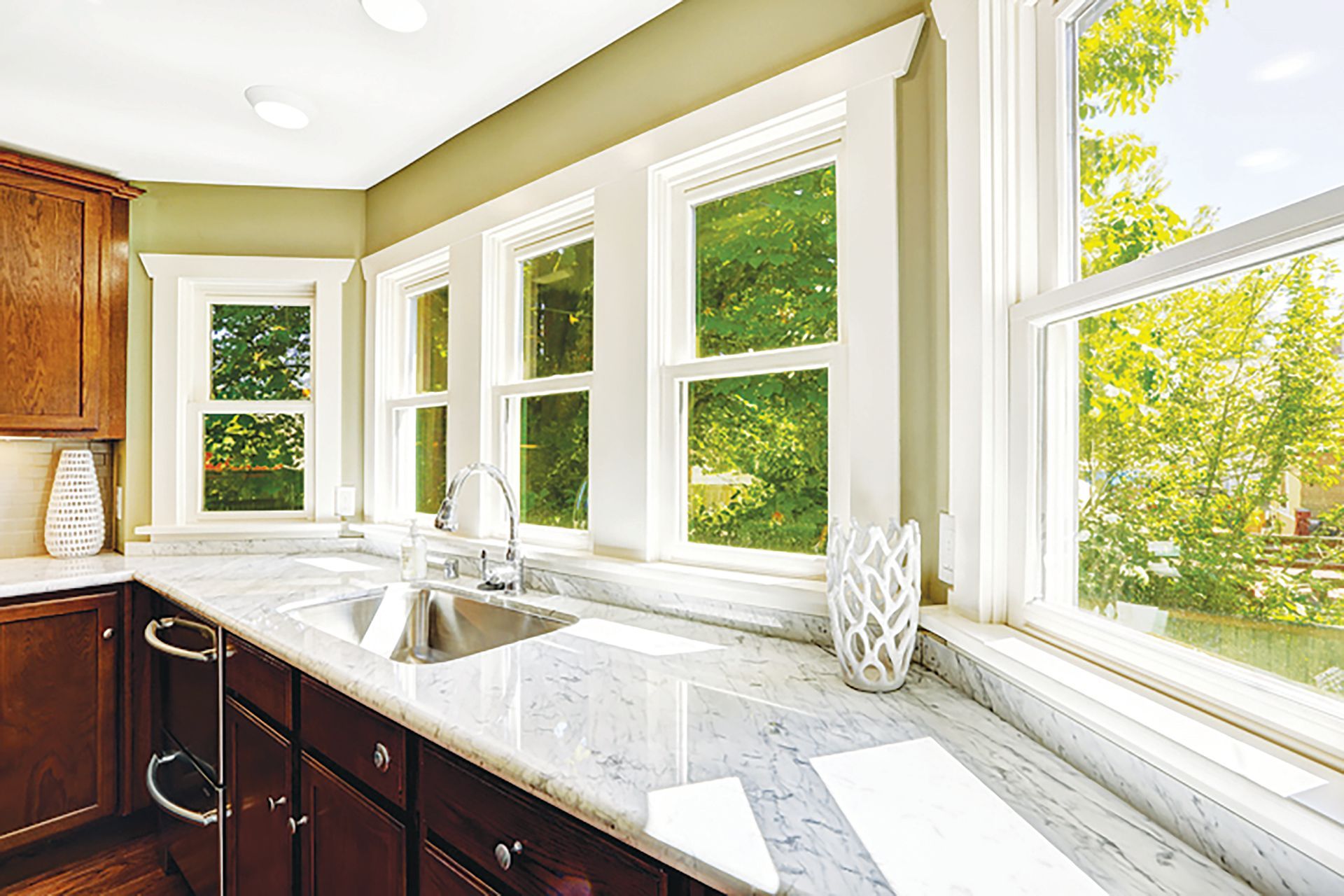 A bright kitchen with dark wood cabinets, marble-patterned countertops, a stainless steel sink, and large sunny windows.