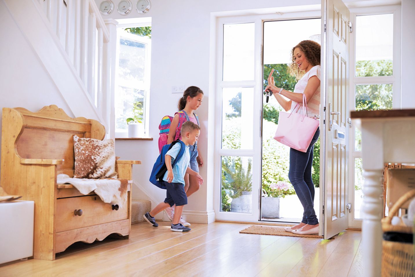 A parent holds a bag and keys as two children wearing backpacks walk through an open front door into a home entryway.