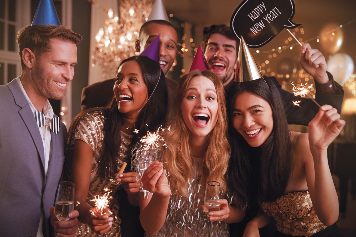 A group of people wearing party hats celebrate with sparklers at a festive New Year’s Eve event.