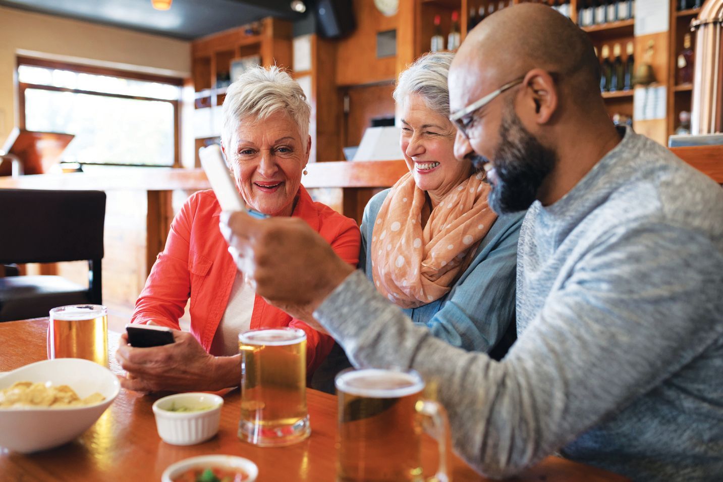 Three people sitting at a table in a bar or pub, smiling while looking at a smartphone held by one person.