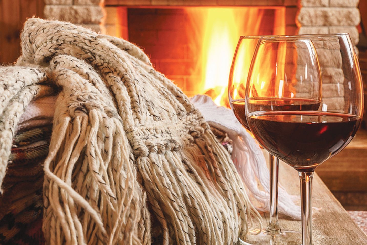 Two glasses of red wine sit next to a cozy, folded knit throw blanket in front of a warm, glowing fireplace.