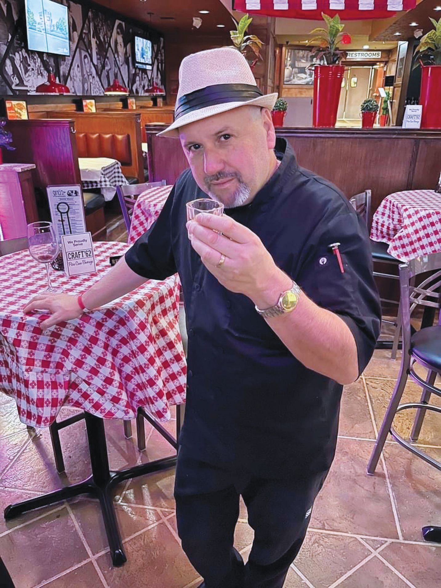 A person in a black chef’s uniform and a fedora toasts with a small glass inside a restaurant with checkered tables.
