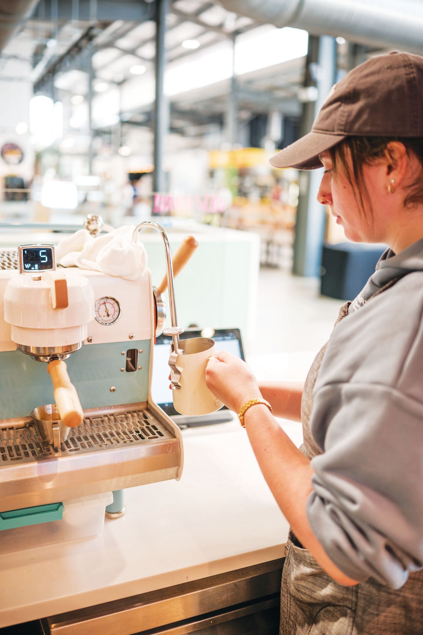 A person in a cap and grey hoodie steams milk using an espresso machine in a cafe.