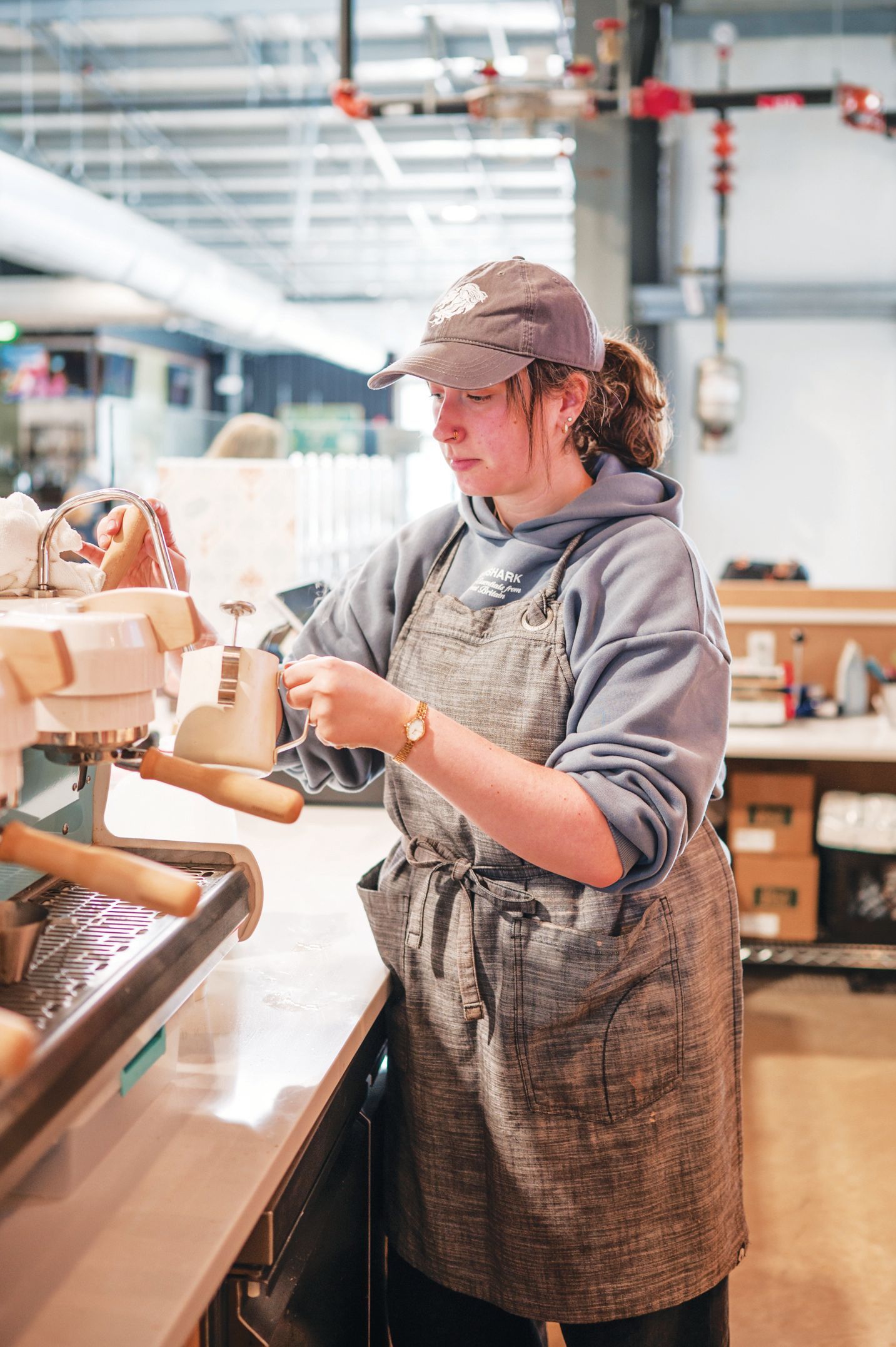 A person wearing a hat and an apron works as a barista, steaming milk with an espresso machine in a cafe setting.