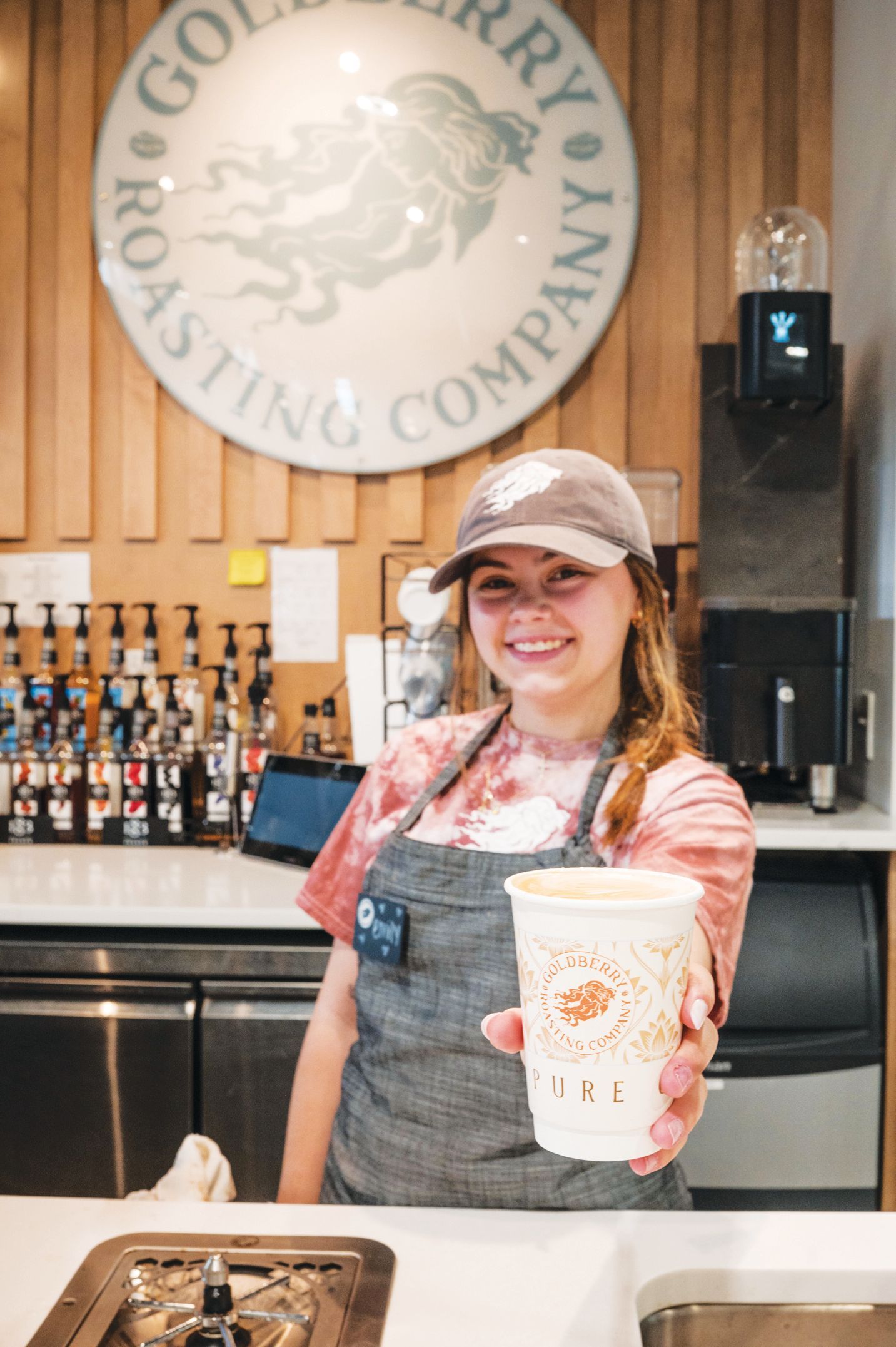 A smiling barista in an apron holding out a branded coffee cup inside Goldberry Roasting Company.