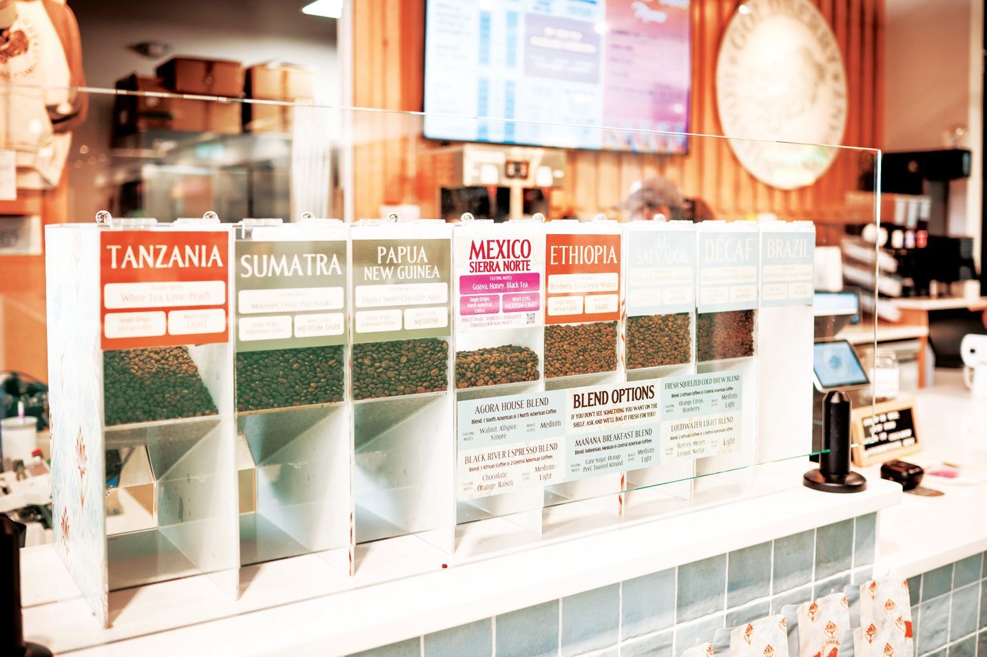 Rows of coffee bean dispensers labeled by origin, including Tanzania, Sumatra, and Ethiopia, in a brightly lit shop.