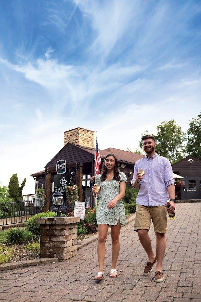 A couple holds glasses while walking along a brick path in front of a rustic building with an American flag under a blue sky.