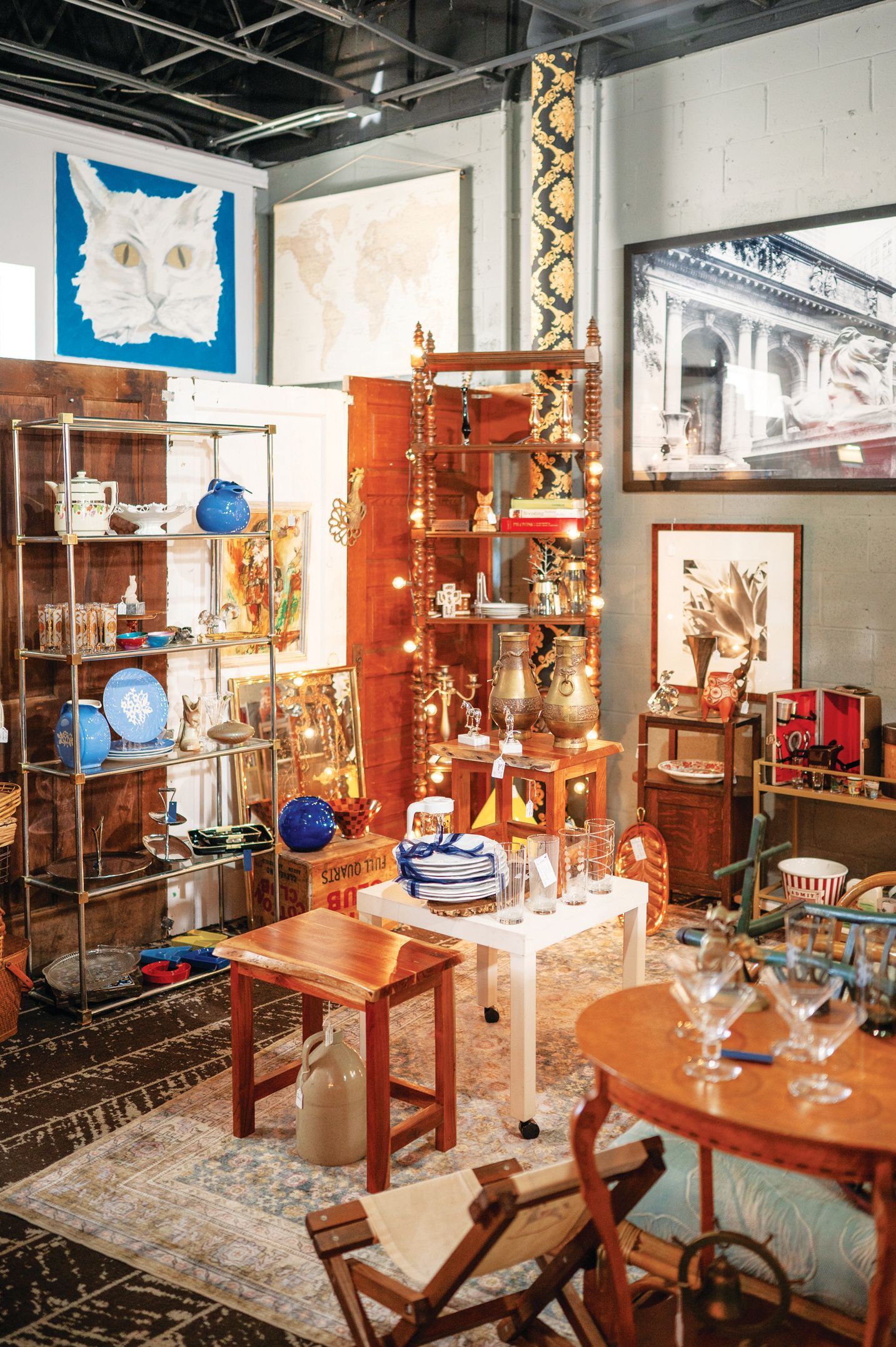 A cluttered antique or vintage store filled with various furniture, decor, artwork, glassware, and a large cat painting.