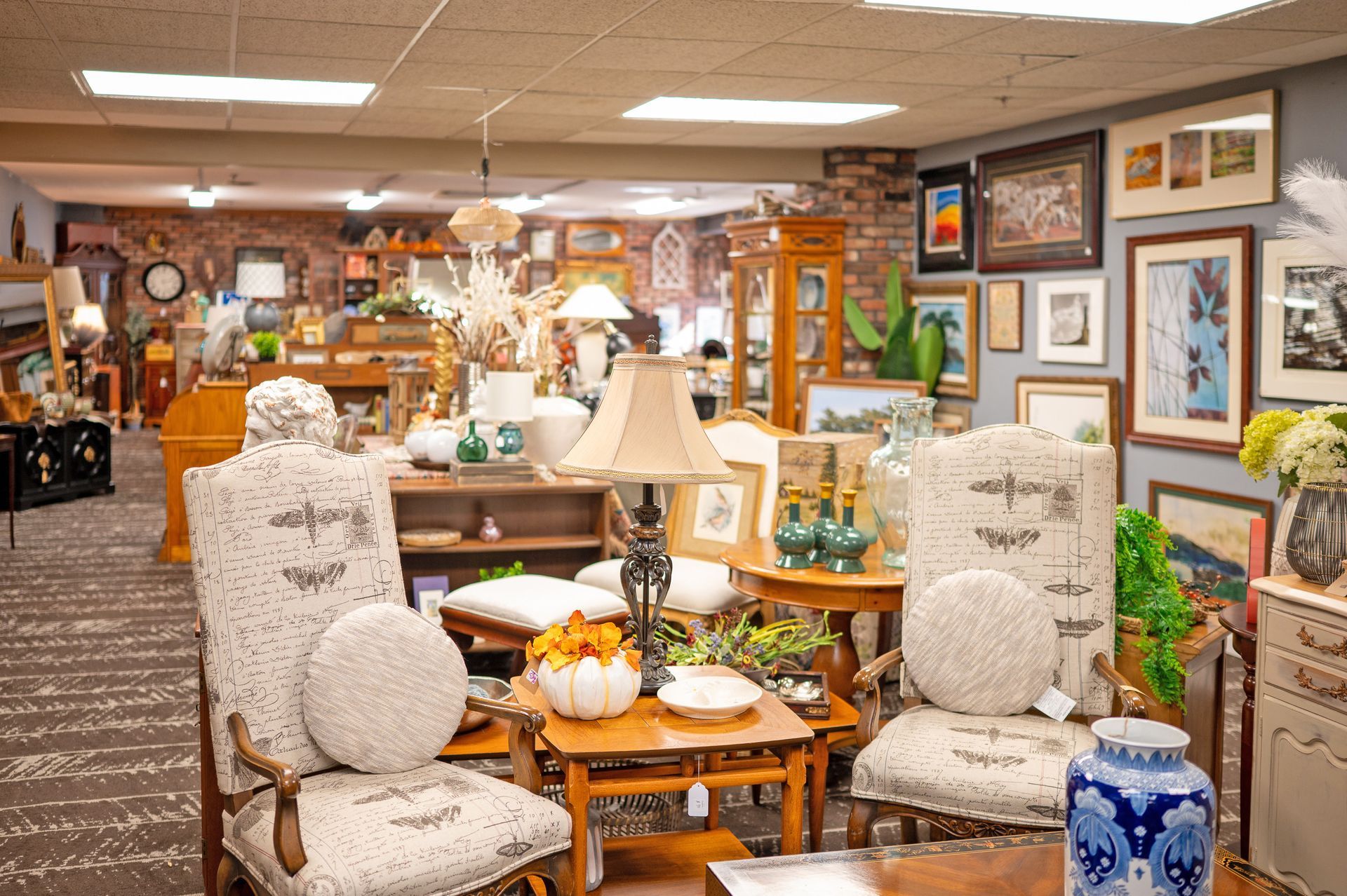 A shop interior featuring two patterned fabric armchairs with round pillows, a central wooden lamp table, and wall art.