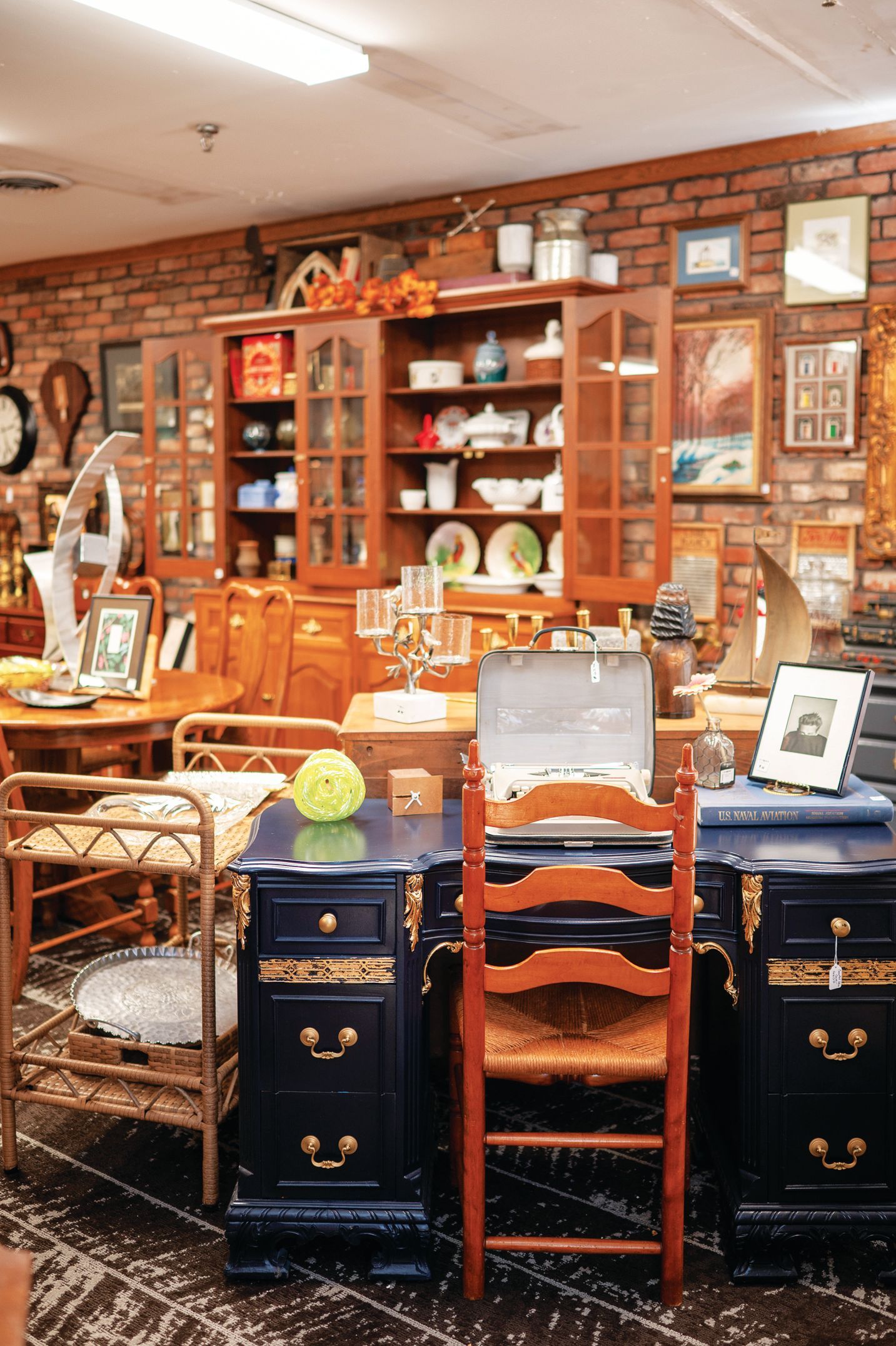 A dark blue, multi-drawer desk with a wooden chair sits in front of a brick wall and a wooden china cabinet in a shop.